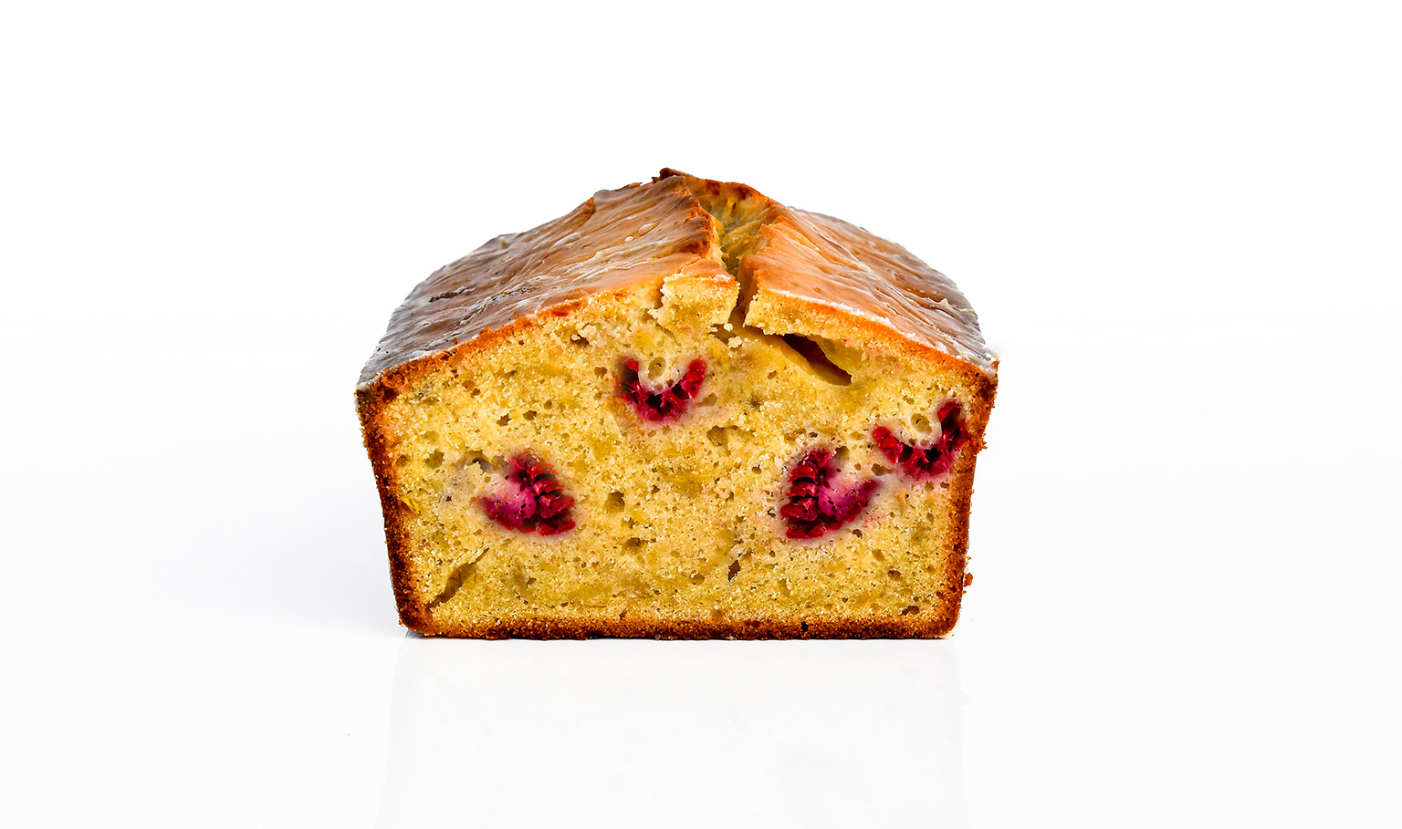 Freshly baked banana pound cake with a golden-brown crust, cut to reveal a moist, buttery interior with visible raspberries. A glossy lime glaze drizzles over the top, pooling slightly at the edges. Plump, fresh raspberries surround the loaf, adding a vibrant pop of red against the warm cake tones.
