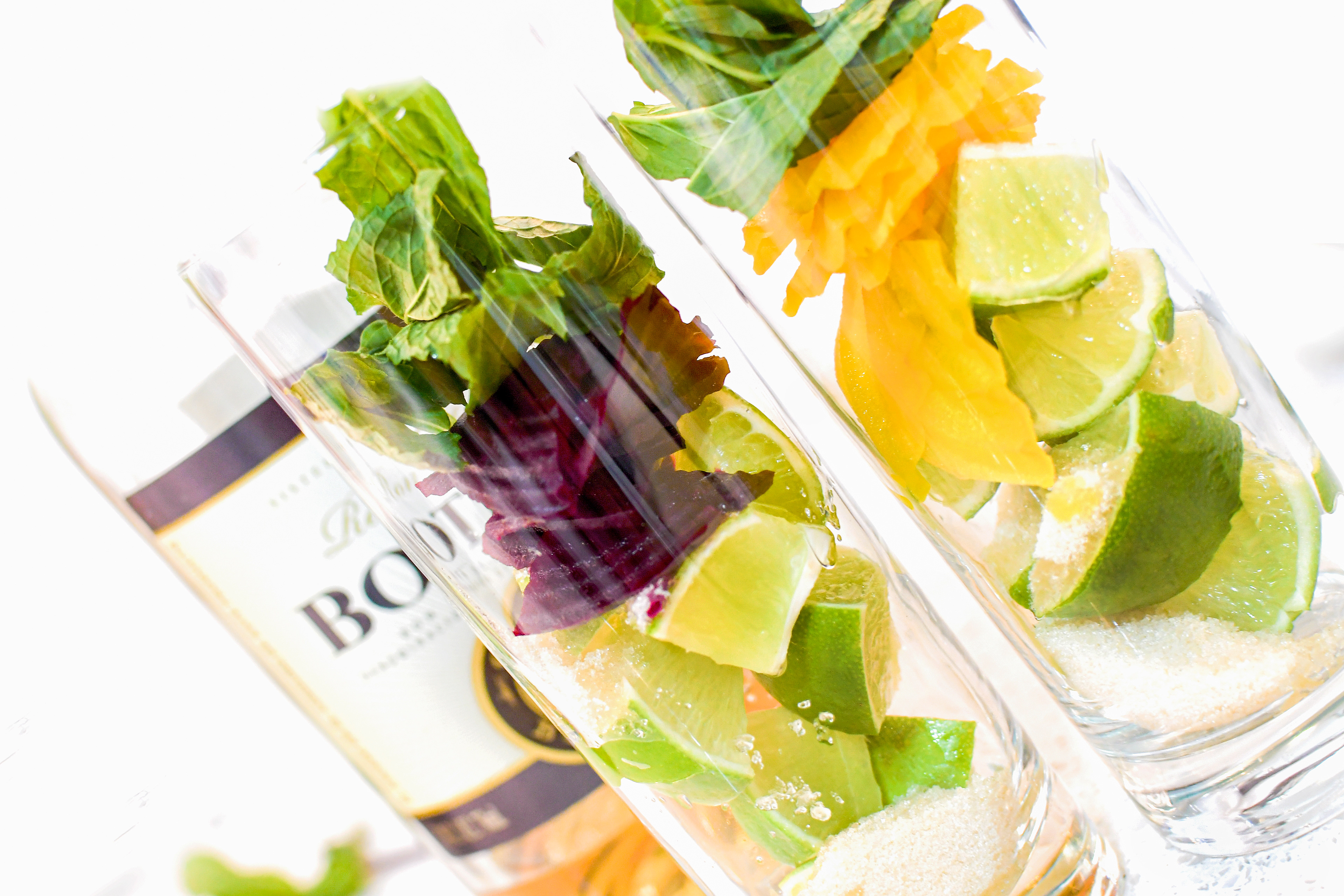 A close-up of two glasses, each containing slices of red and yellow beets, cut limes, crystalline sugar, and fresh mint leaves. Both glasses are set against the backdrop of a bottle of Ron Botran Anejo 8-Years rum. The image showcases the fully prepared ingredients for making Beet Mojito, with the addition of mint leaves, illustrating the next step in the cocktail preparation.