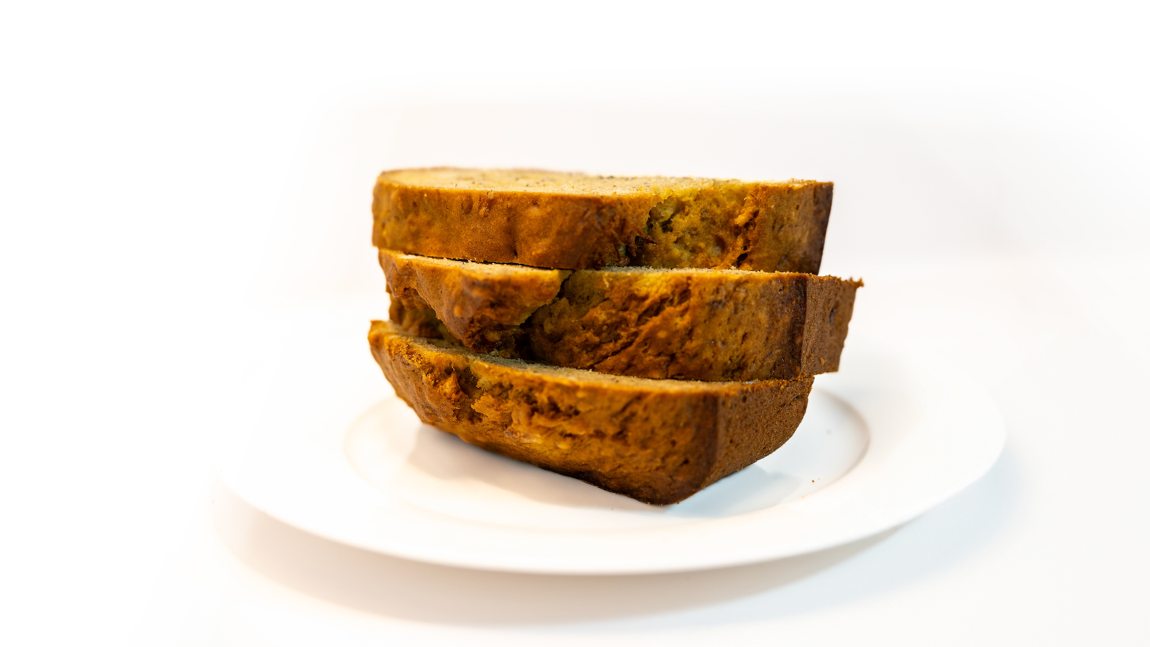 Three evenly sliced pieces of moist banana bread arranged on a simple white plate, set against a plain white background. The bread's golden-brown crust contrasts beautifully with its soft, tender interior, revealing flecks of banana and optional add-ins like walnuts or chocolate chips.