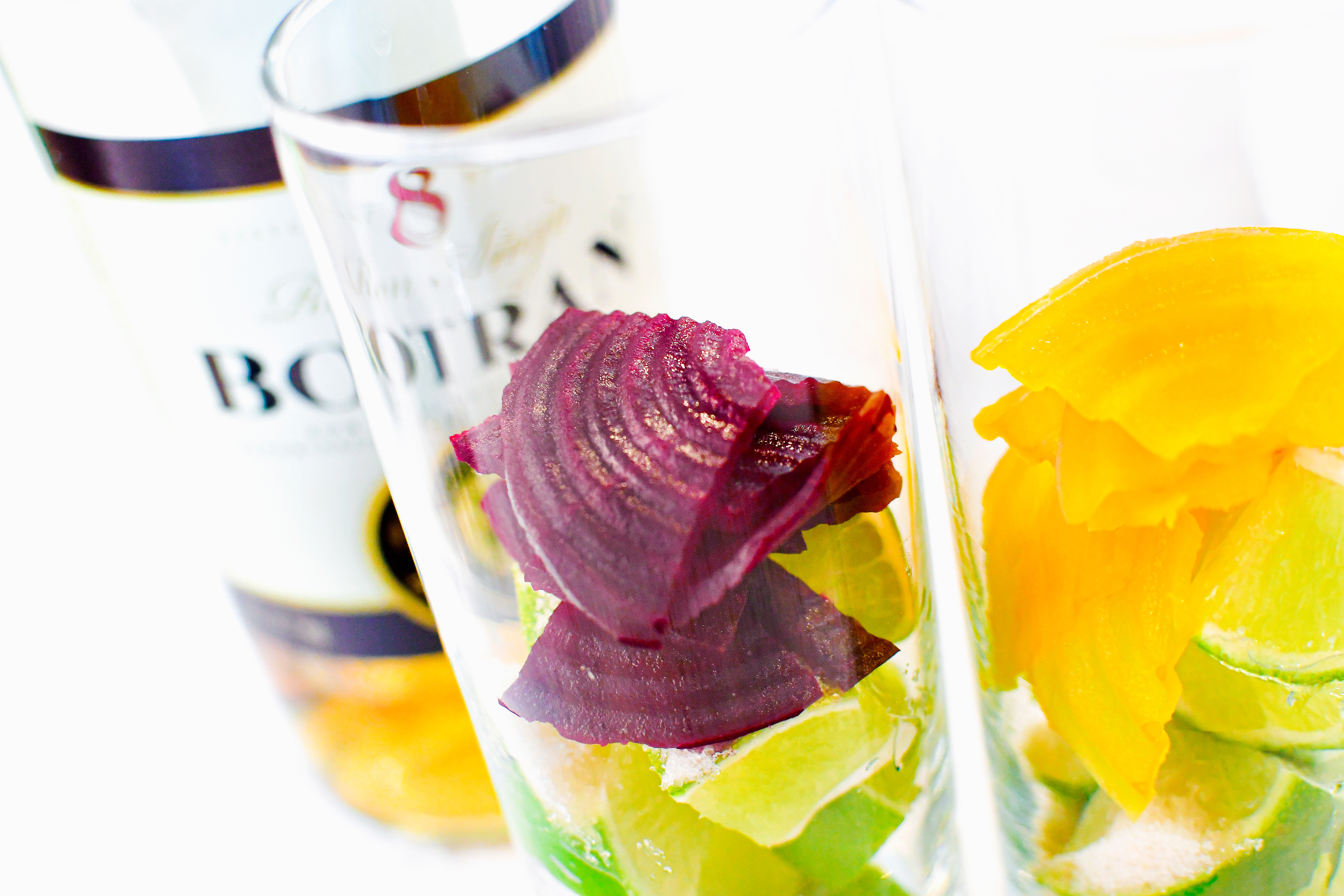 A close-up of two glasses, each filled with slices of red and yellow beets, respectively. Both glasses also contain cut limes and crystalline sugar. A bottle of Ron Botran Anejo 8-Years rum is visible in the background. The image shows the prepared ingredients, illustrating the next step in making the Beet Mojito, with all components ready for mixing.