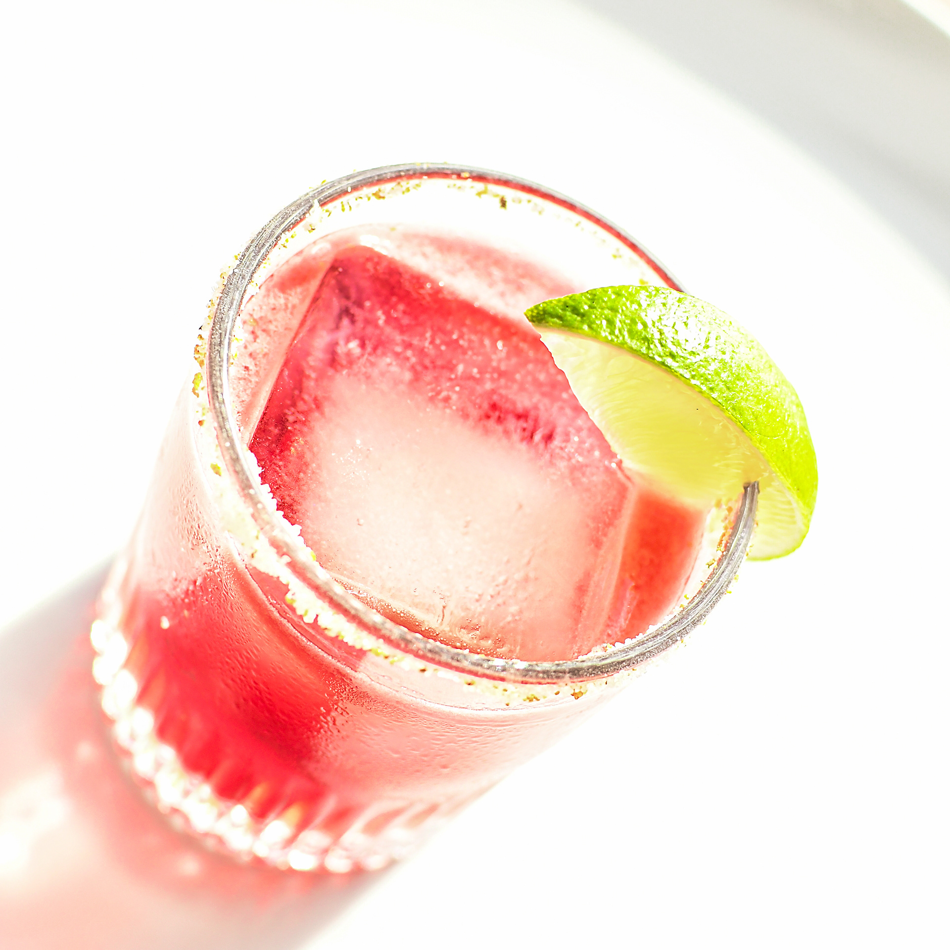 A stylish Hibiscus Margarita on a white background, showcasing its vibrant, deep color. The glass, garnished with a lime wedge and featuring a salted rim, is filled with a large ice cube, which helps keep the drink chilled without diluting it quickly. The cocktail, made with tequila blanco, hibiscus tea, freshly squeezed lime juice, and agave syrup, highlights the dramatic and rich hues of the hibiscus, creating a refreshing and nutrient-rich summer drink.
