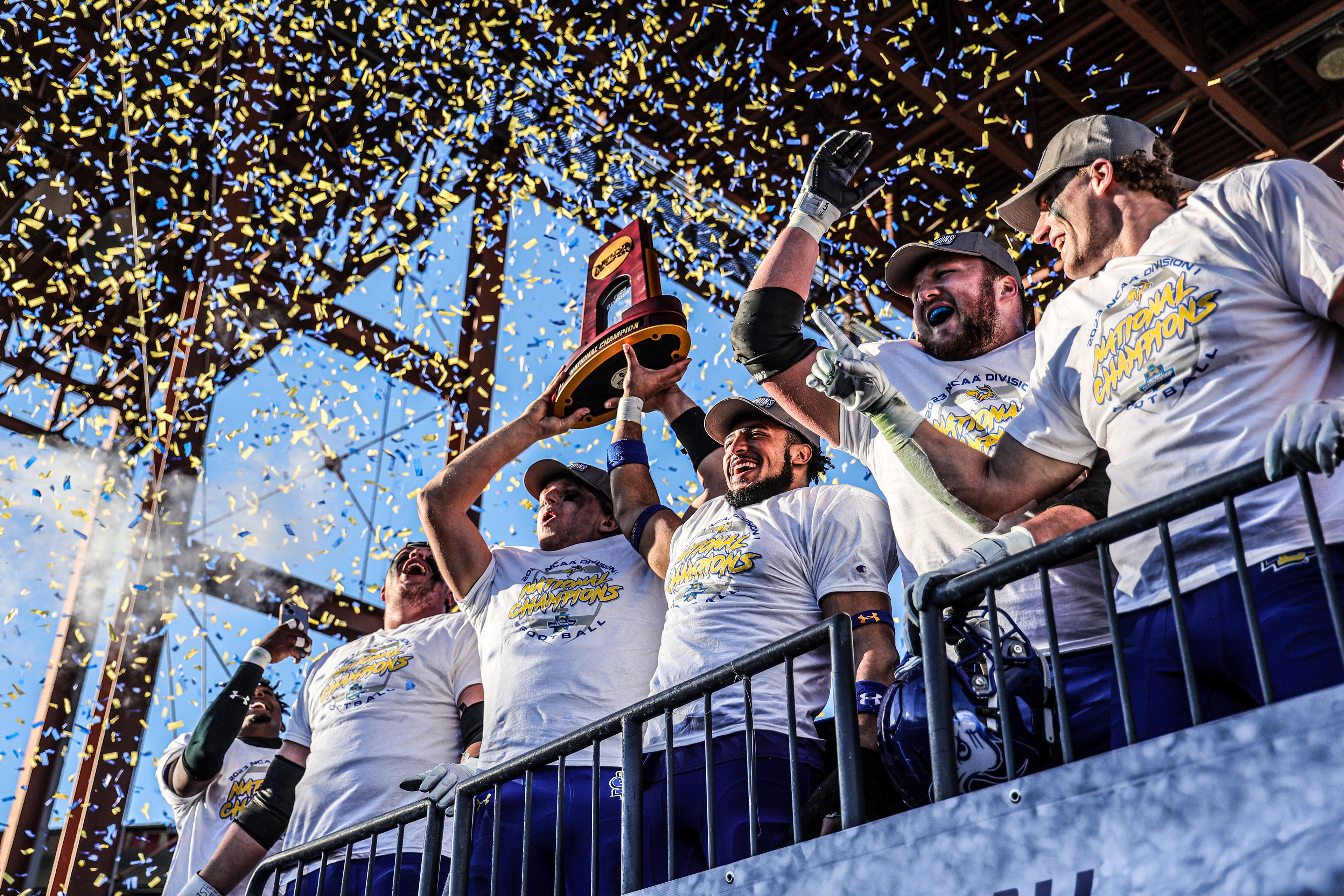 South Dakota State players hoist the NCAA trophy as blue and yellow confetti rains down after defeating Montana in the 2023 FCS National Championship in Frisco, Texas. The win capped an undefeated season and secured back-to-back national titles for the Jackrabbits.