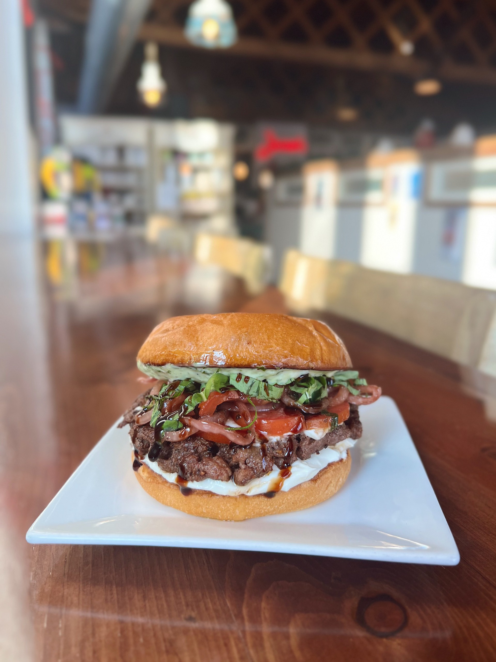 Capo Caprese Burger - Josiah's Coffeehouse - A flavorful fusion of Italian and classic American: A toasted brioche bun stacked with our garlic ricotta cream cheese spread, a 6oz grilled patty, house made tomato-onion bruschetta and basil, balsamic drizzle, and our fresh pesto aioli.