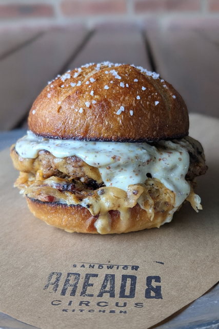 Das Brat! - Bread & Circus - A 6oz house-made bratwurst patty topped with rich beer cheese, house sauerkraut braised with beer, bacon, and onions, house grainaise, all served on a custom Breadico pretzel bun — inspired by classic German biergarten flavors.