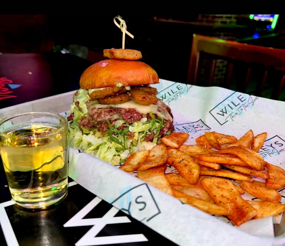 The Pickleback Smash - Wileys Bar - Introducing The Pickleback Smash! Two smash patties, melted dill Havarti, bacon jam, pickled lettuce slaw, loaded with hot honey fried pickles dripping in house dill sauce served on a toasted brioche bun. It’s briny. It’s crispy. It’s sweet perfection. One bite – Full Pickleback Energy. Every burger served with a mini size Pickle Beer.