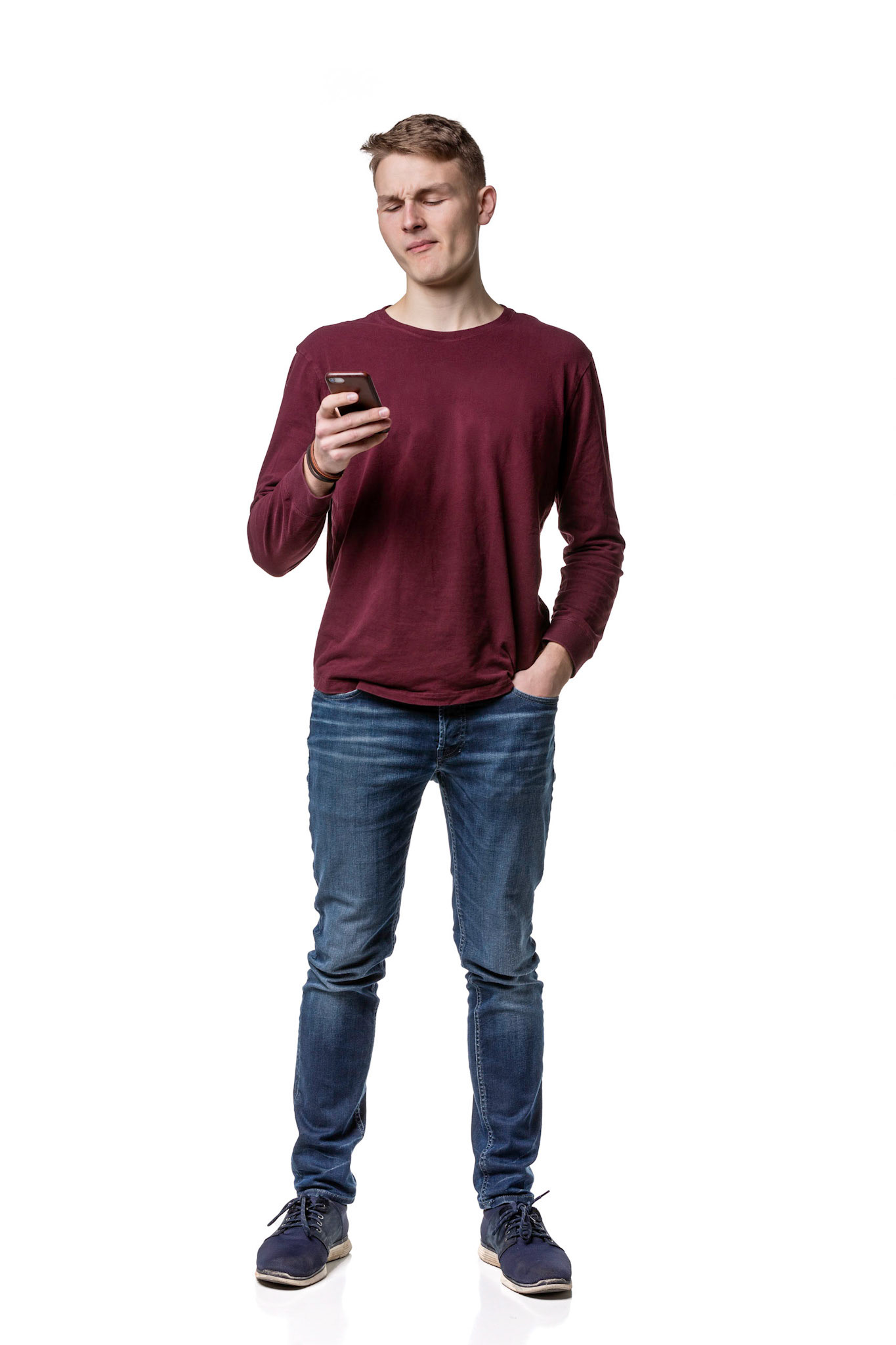 A young man wearing jeans and casual maroon shirt holds a phone isolated on a white background