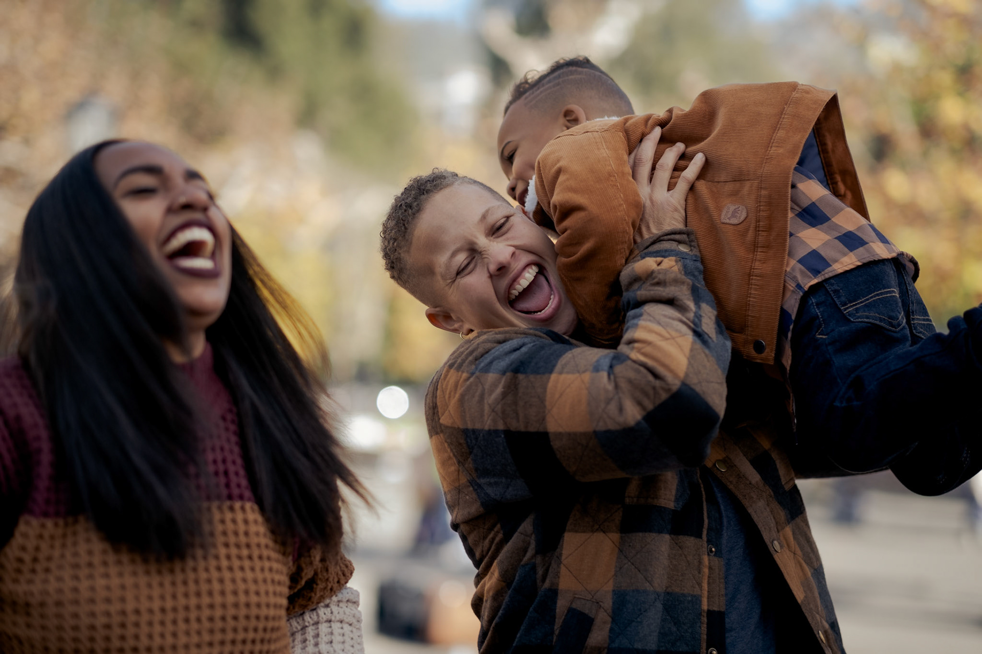 there are three people. on the left, a woman wears a knit sweater and stands next to her wife, who wears a flannel shirt and holds up their three year old son who is wears a matching flannel shirt. both mothers appear joyful, laughing with their eyes closed and mouths open in a moment of tremendous happiness. their son's expression is barely visible, but he is also laughing as he "flies" through the air in his mother's strong arms.