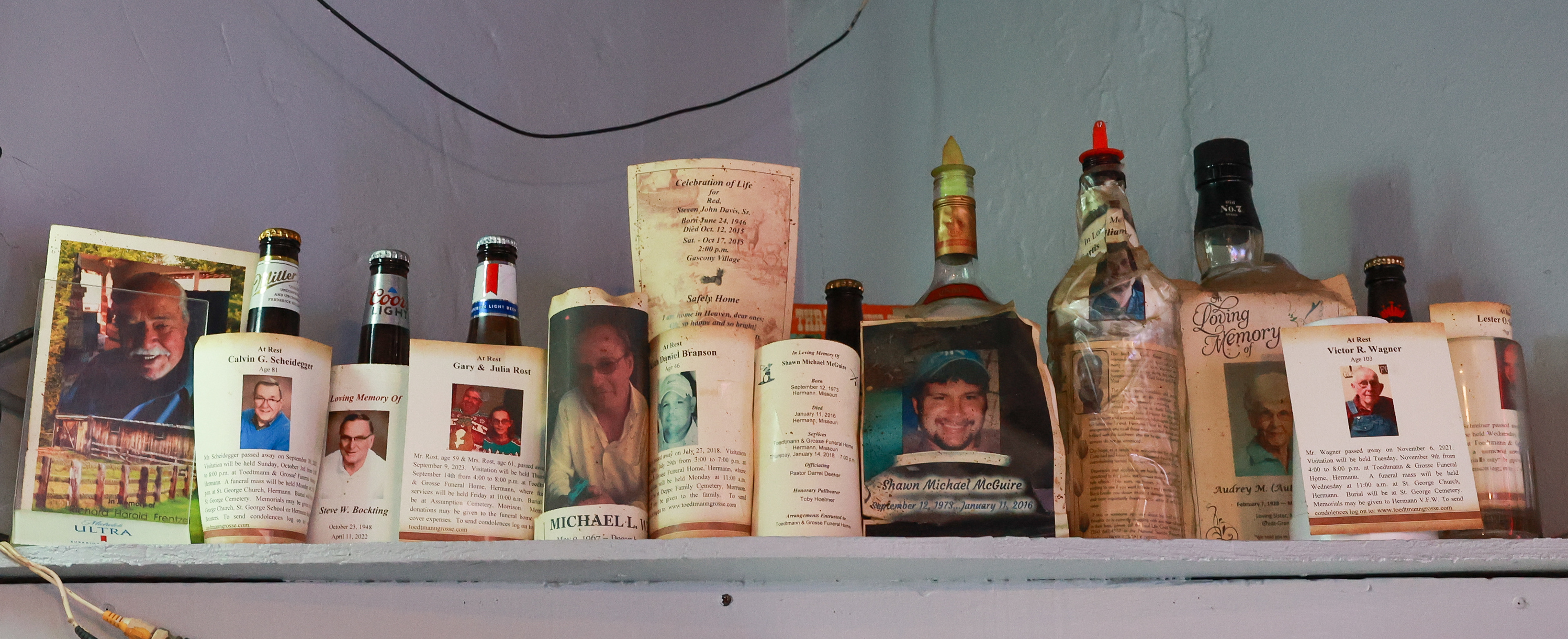 A collection of obituaries for the tavern’s regular customers who have passed sit on a shelf on Tuesday, March 5, 2024 at Sharp Corner Tavern in Hermann, Mo. The memorial photos are taped to empty bottles of the customers’ drink of choice when they used to frequent the tavern.