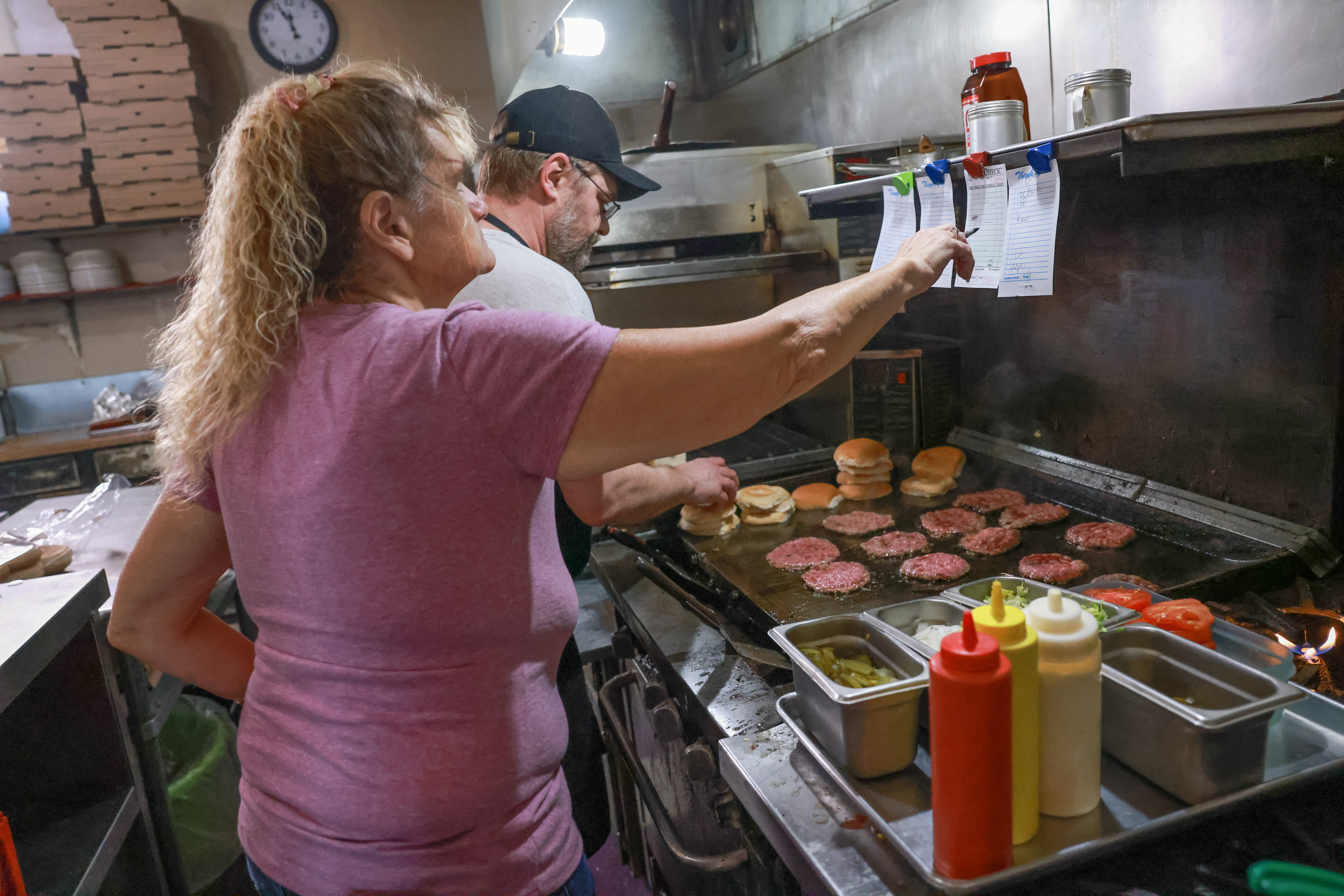 Toby Hoelmer cooks Herbie burgers while waitress Jackie Rost delivers another order on Tuesday, March 12, 2024, at the Sharp Corner Tavern in Hermann, Mo. The Tavern is best known for their “Herbie burgers”, greasy smash burgers named after a previous owner Herb Mueller. Beyond the burgers, the Tavern serves daily specials, such as meatloaf and fried fish sandwiches.