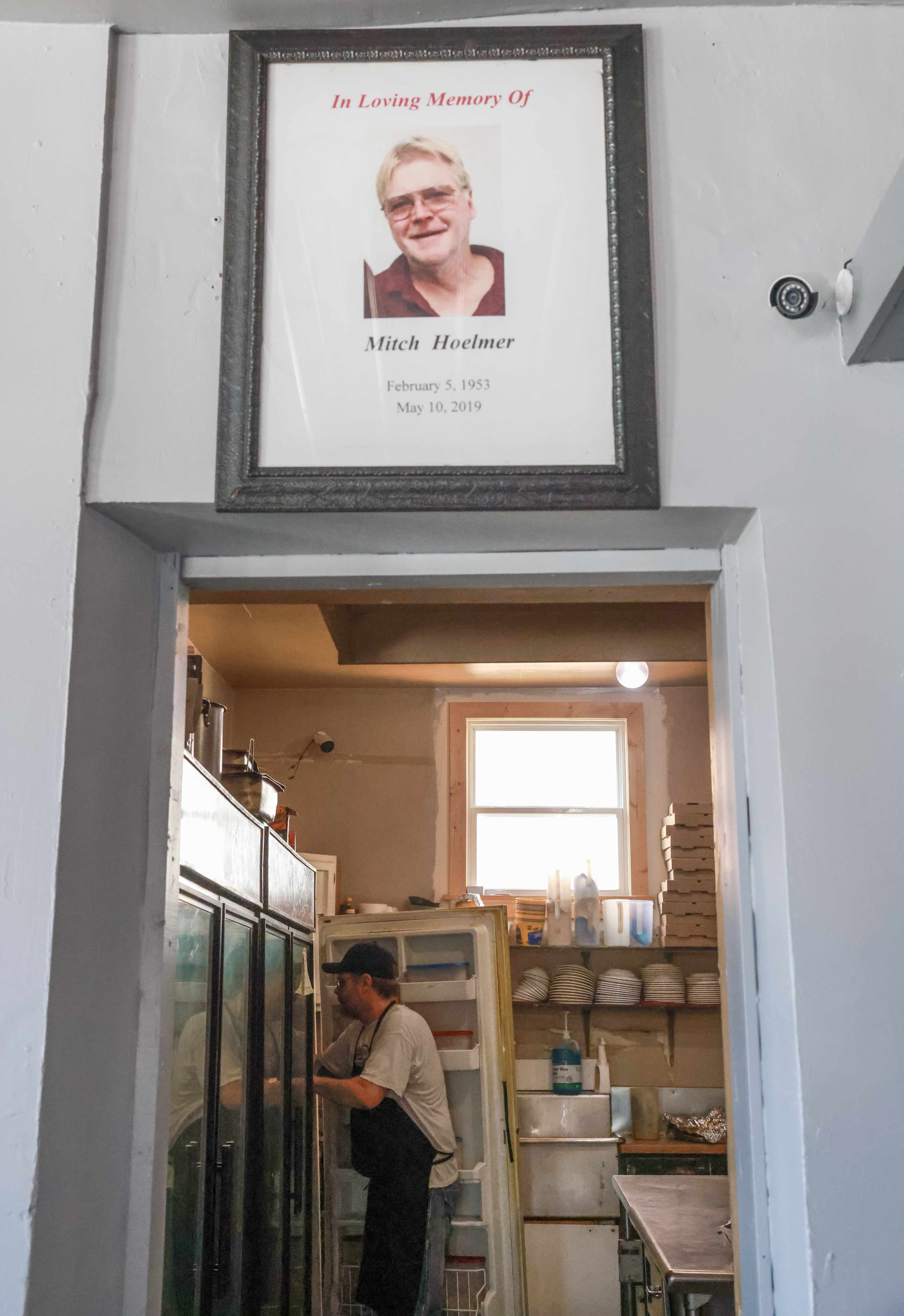 Toby Hoelmer gathers ingredients for the lunch rush under a memorial poster for his father Mitch Hoelmer on Tuesday, March 12, 2024, at the Sharp Corner Tavern in Hermann, Mo. Toby and Sarah, who has previously married to Mitch, have co-owned the bar since his passing in 2019.