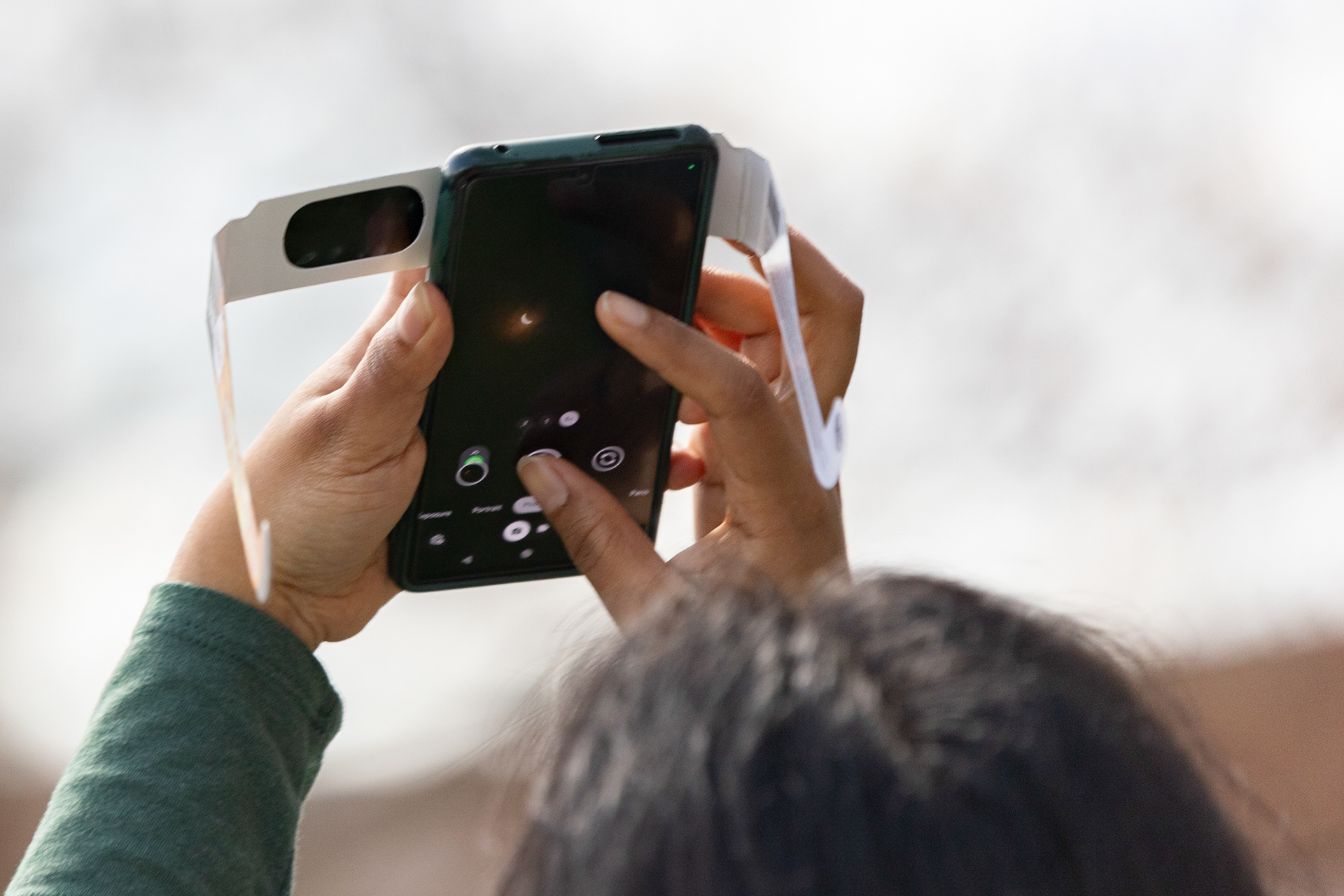 A person uses their solar eclipse glasses to take a picture of the eclipse on their phone during peak viewing at Paw Print Park, Ohio University, Athens, April 8, 2024.