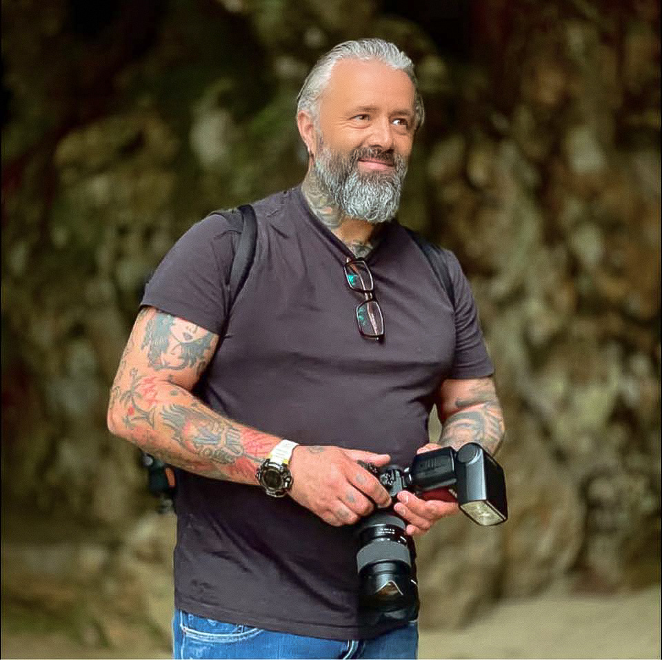 Luis Rocha, fotógrafo profissional no Algarve, posando com câmara DSLR e flash externo num cenário natural de folhagem