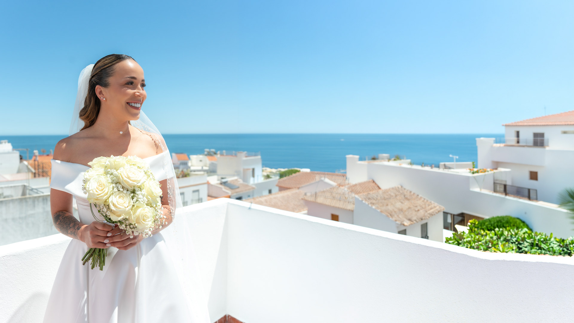 Fotografia documental de casamento em Albufeira: noiva Sara sorridente no terraço durante os preparativos, segurando um bouquet de rosas brancas com o mar azul e as casas típicas do Algarve em fundo. Documentary wedding photography in Albufeira: bride Sara smiling on a terrace during preparations, holding a white rose bouquet with the blue ocean and typical Algarve white houses in the background.