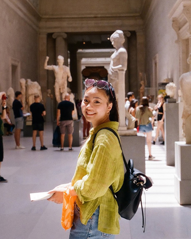 Sarah Nuzum stands in the middle of the image, smiling at the camera. She is standing in the middle of an art gallery.