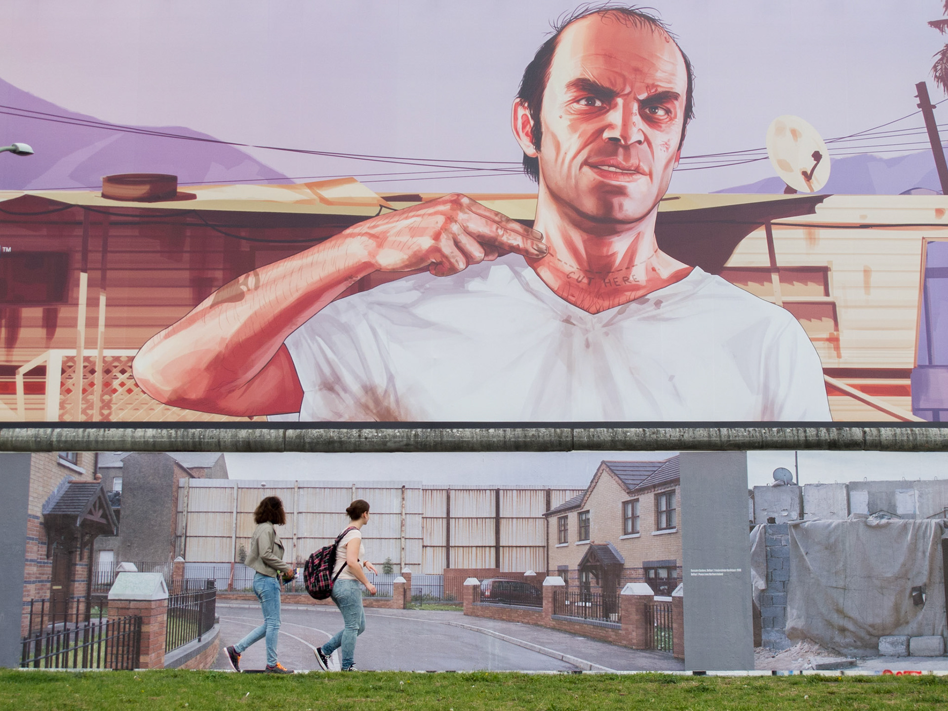 Tourists visiting the East Side Gallery in Berlin are looking at the Peace Lines in Belfast, a photograph by Kai Wiedenhöfer and part of his WallOnWall project; a photo exhibition on the Berlin Wall about walls that separate people worldwide. Behind the Berlin Wall, a large Grand Theft Auto V video game advertisement is shielding yet another redevelopment project along the Mühlenstraße
