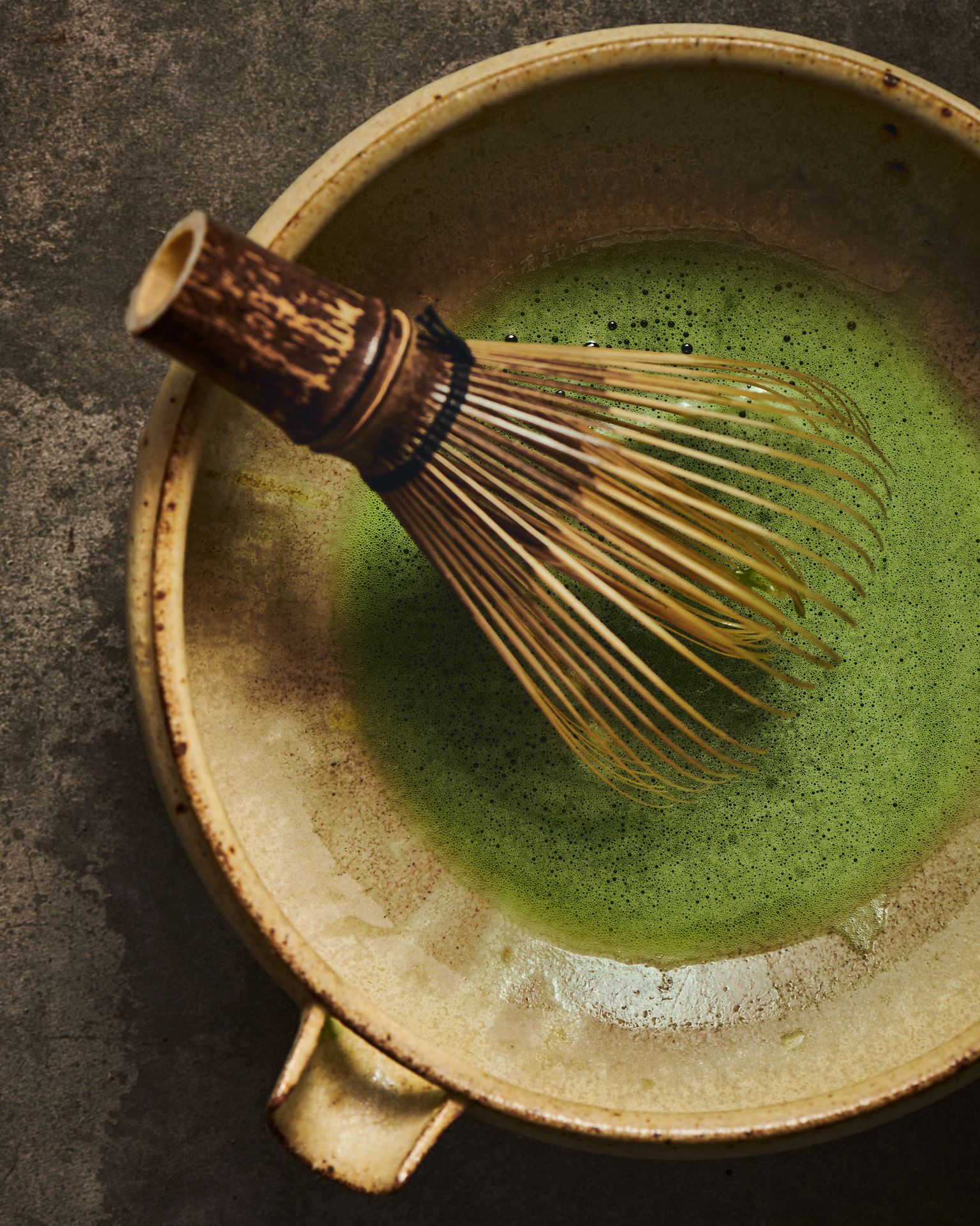 Kata Endrodi Montreal food and product photographer, detailed shot of frothy matcha tea in a textured ceramic bowl, showcasing the delicate bubbles of a traditional whisking technique and organic tones.