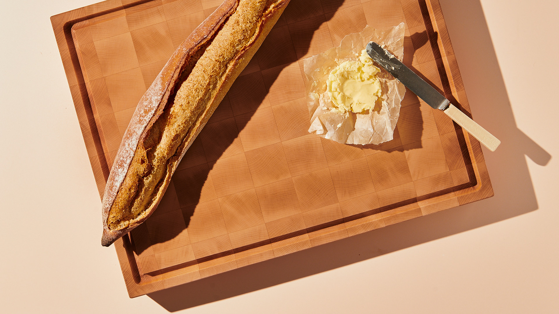 Vue en surplomb d'une baguette artisanale et de beurre frais sur une planche à découper en bois de bout sous un éclairage directif, par Kata Endrodi, photographe culinaire à Montréal.