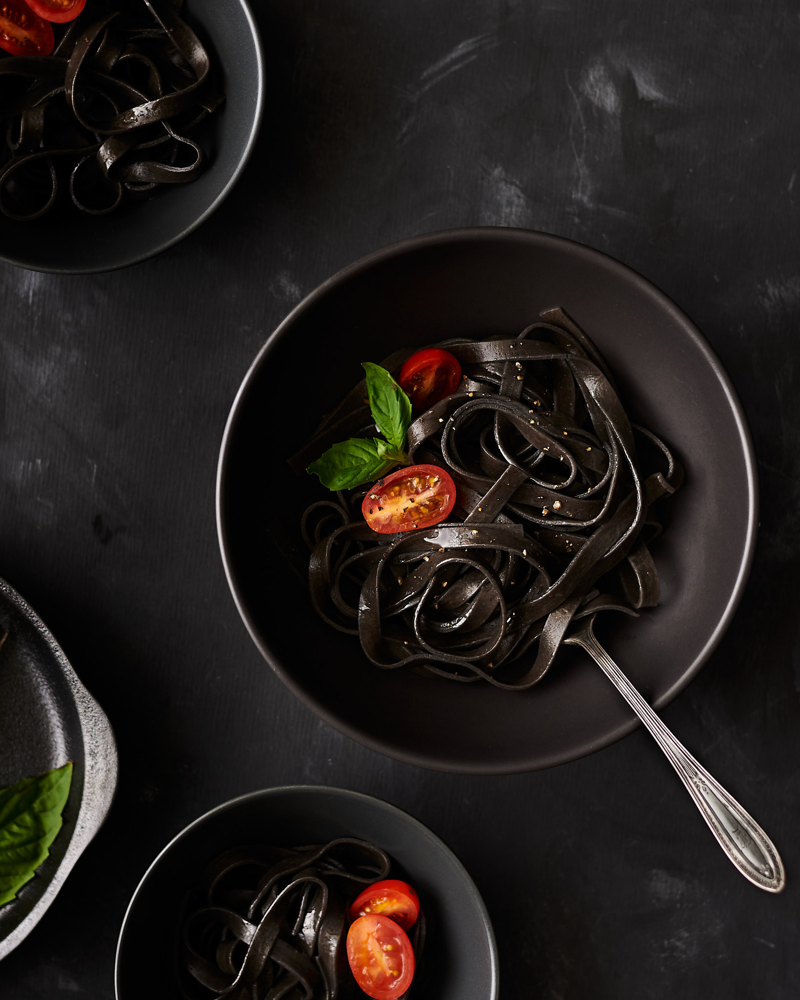 A food photography black squid ink pasta in black bowls image with tomato and basil, made by Kata Endrodi, a food photographer in Montreal
