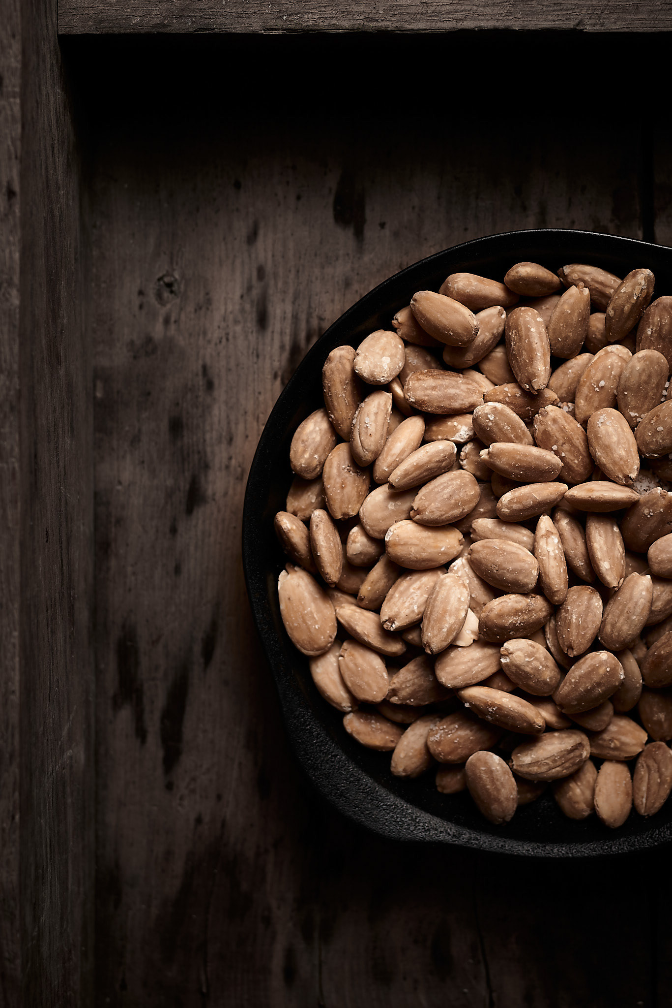 A food photography salted roasted almonds on black plate and aged wood tray image, made by Kata Endrodi, a food photographer in Montreal