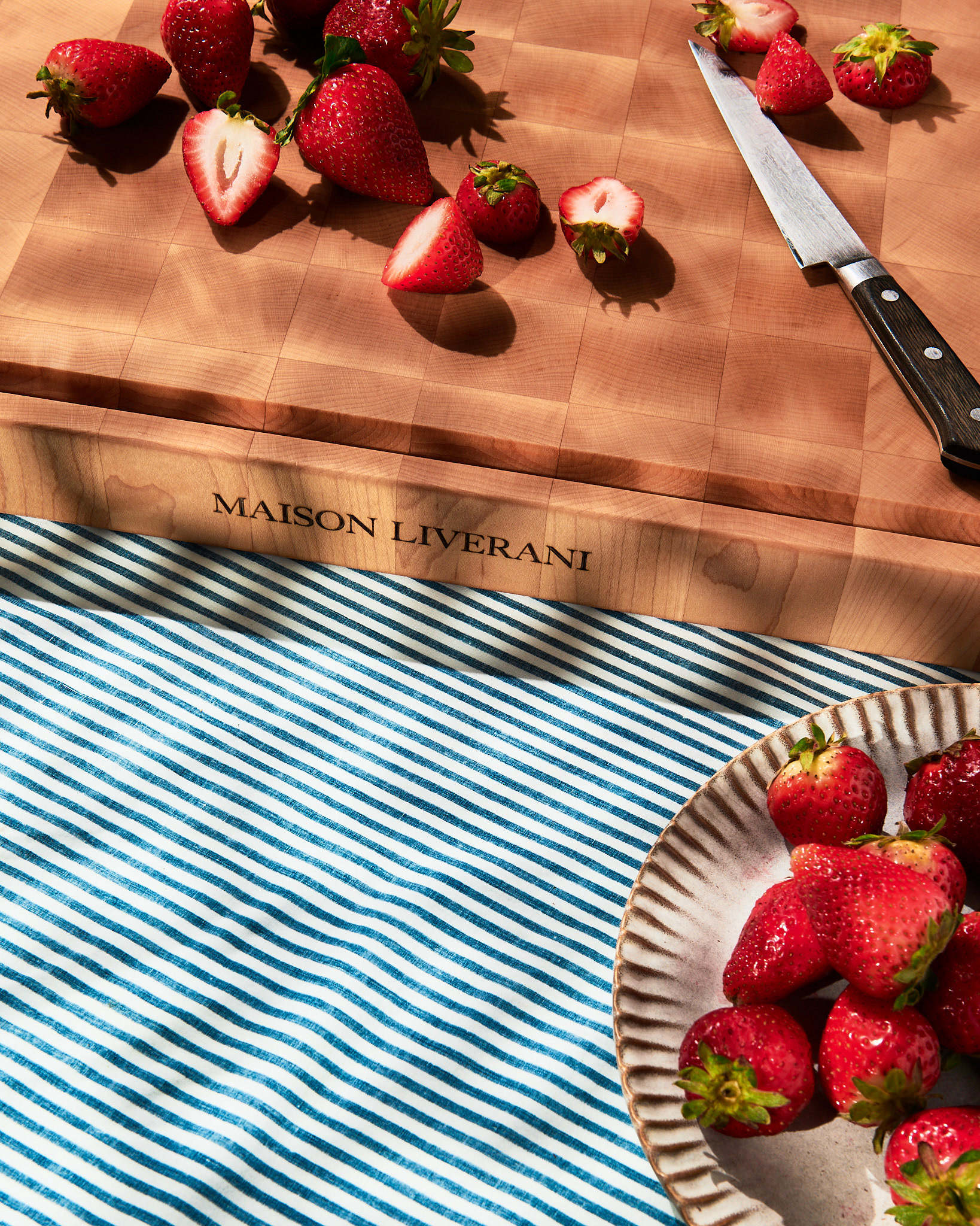 Close-up view of fresh strawberries and a chef knife on an end-grain wooden cutting board in warm sunlight, by Kata Endrodi, food photographer in Montreal.