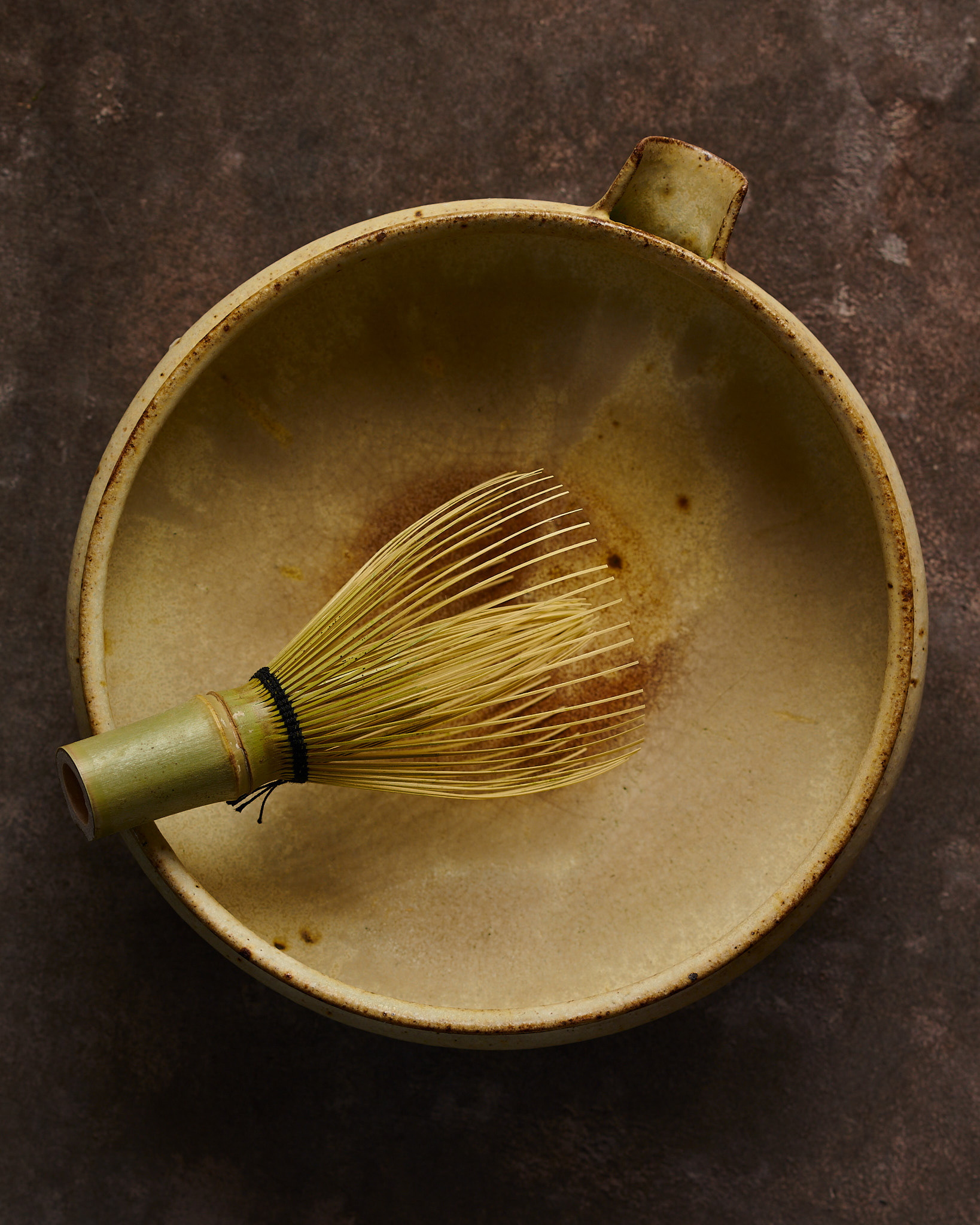 Kata Endrodi Montreal food and product photographer, lifestyle photography featuring a handmade yellow ceramic matcha bowl and whisk on a rustic brown background