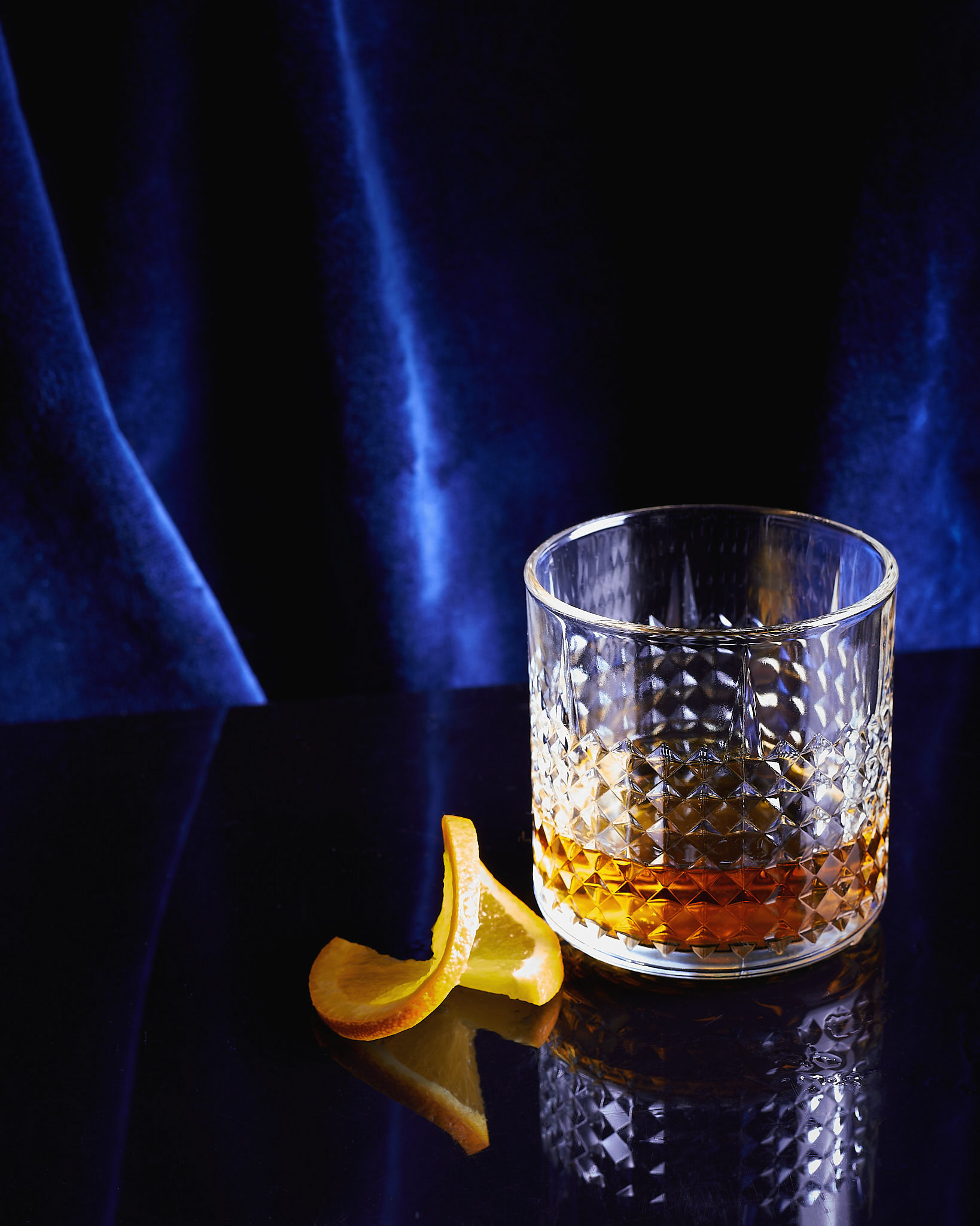 A food photography glass of whiskey image with blue velvet background and a slice of orange on the side, made by Kata Endrodi, a food photographer in Montreal
