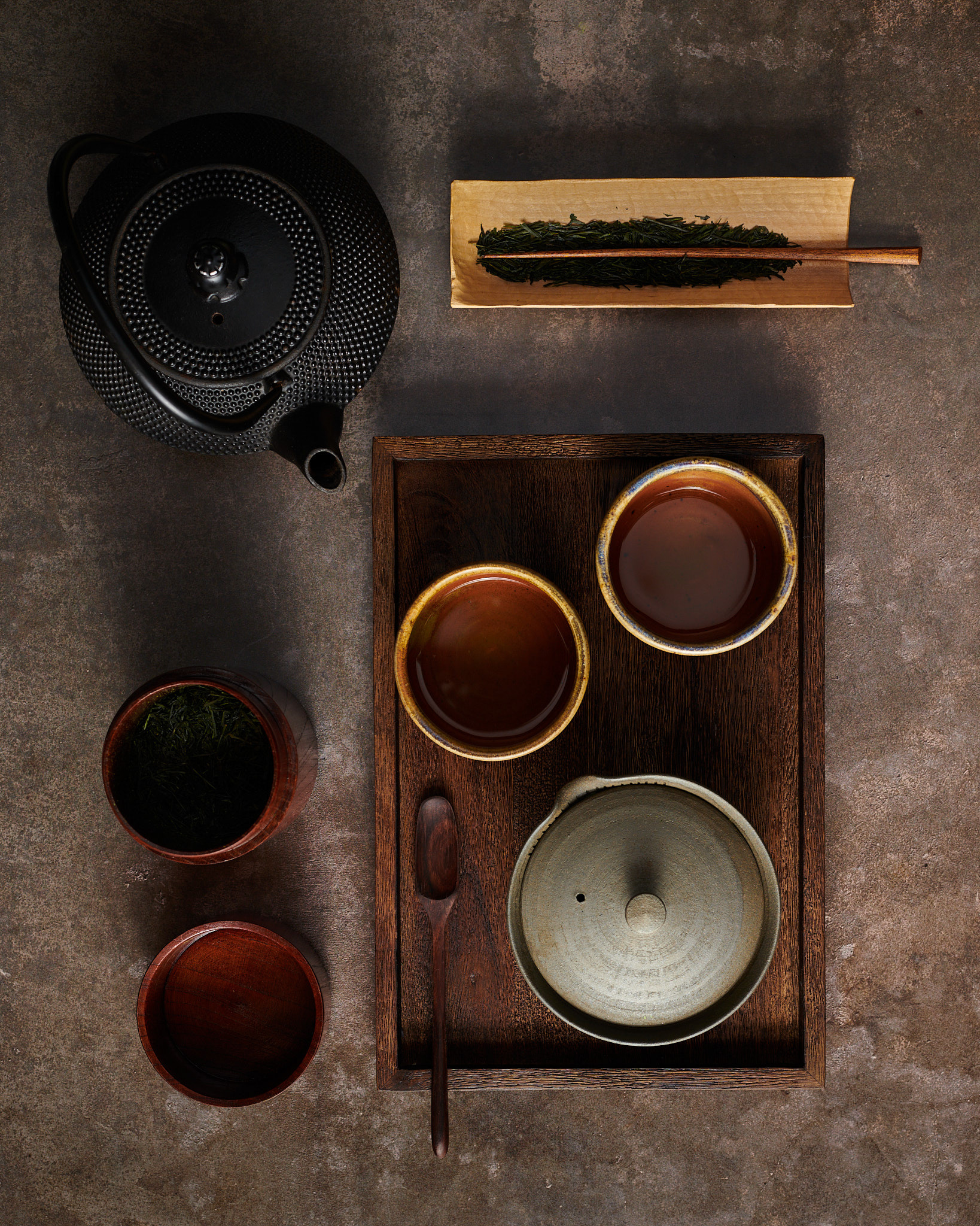 Kata Endrodi Montreal food and product photographer, lifestyle photography featuring a cast iron black teapot, loose leaf Japanese tea in wooden container, loose leaf tea holder, two ceramic bowls and a small teapot on a dark wooden tray on a brown surface.