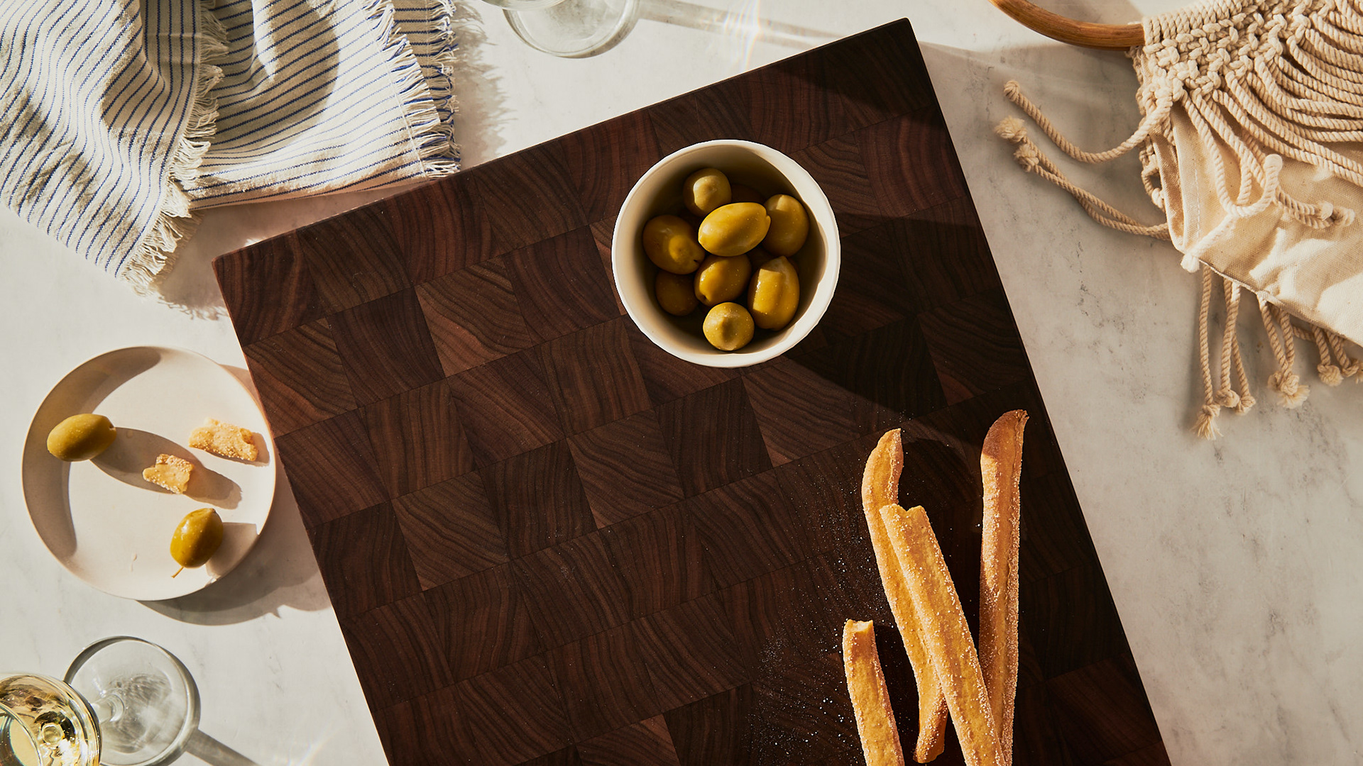 Vue en surplomb style de vie d'une planche à découper en bois de bout foncé garnie d'olives, de grissinis, d’une serviette en lin et d’un sac en macramé, par Kata Endrodi, photographe lifestyle à Montréal.