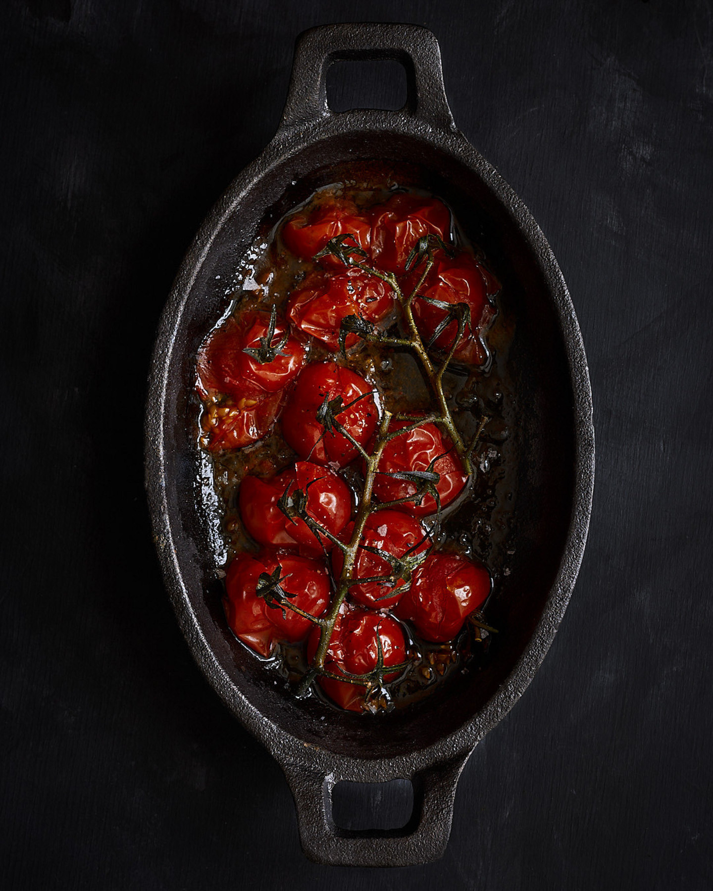 A food photography roasted cherry tomatoes image in cast iron skillet on dark backdrop, made by Kata Endrodi, a food photographer in Montreal
