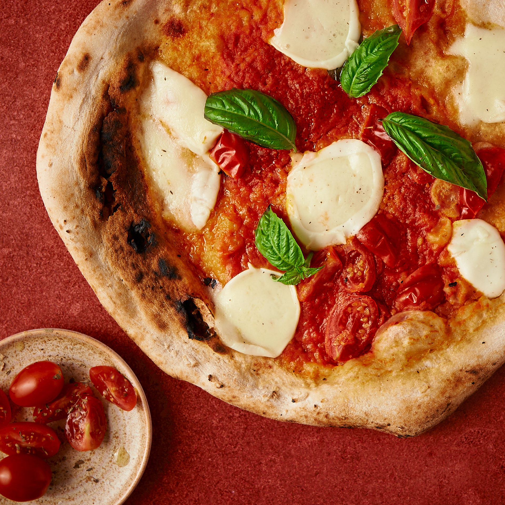 Artisanal baked pizza photography with fresh basil and tomatoes on a textured red background, art directed by Montreal food photographer Kata Endrodi.