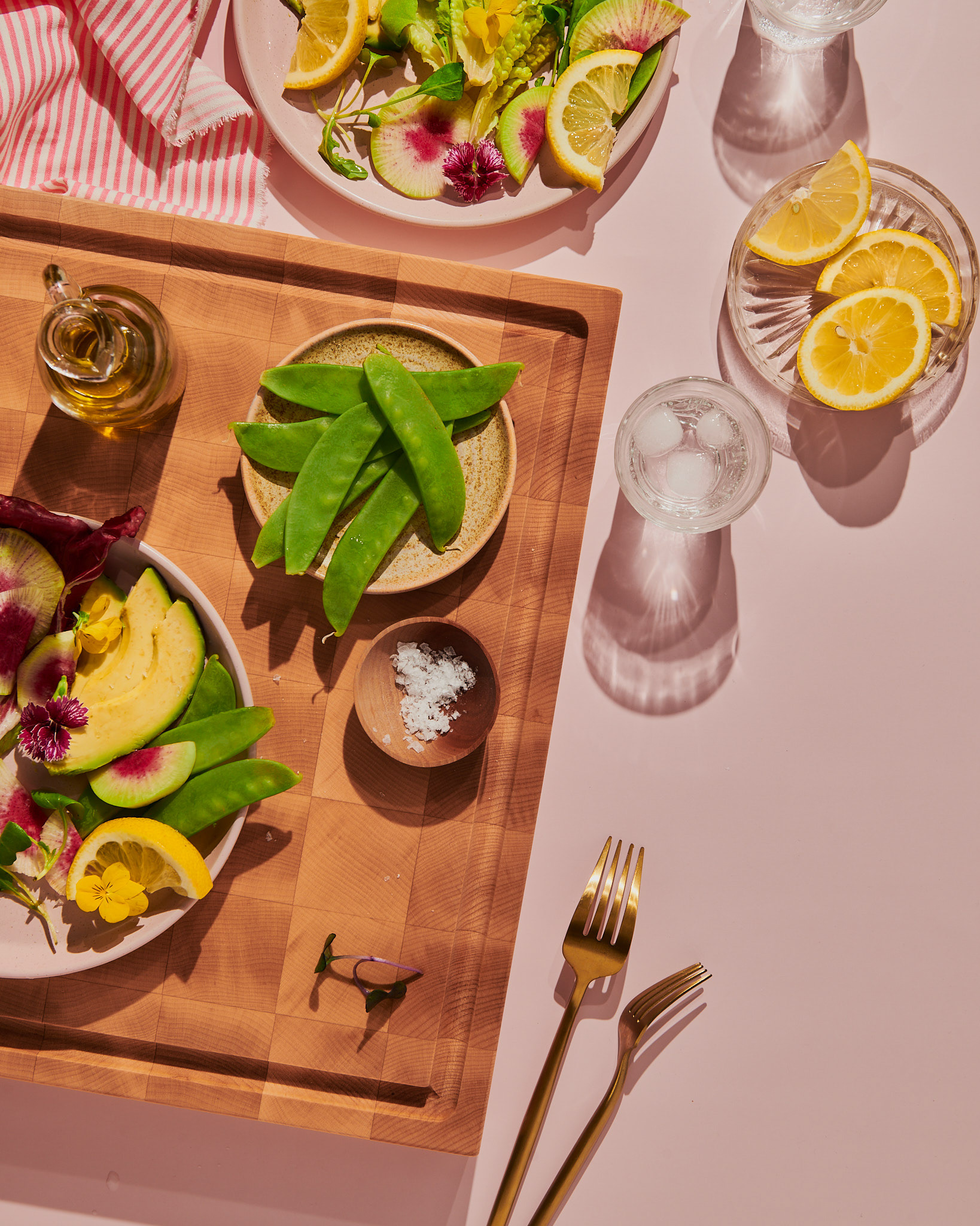 Overhead flat-lay lifestyle food photography by Kata Endrodi in Montreal, featuring a bright dining table with hard shadows, a wooden cutting board with fresh snap peas, an avocado salad with edible flowers, sliced lemons, gold forks, and a glass of water.