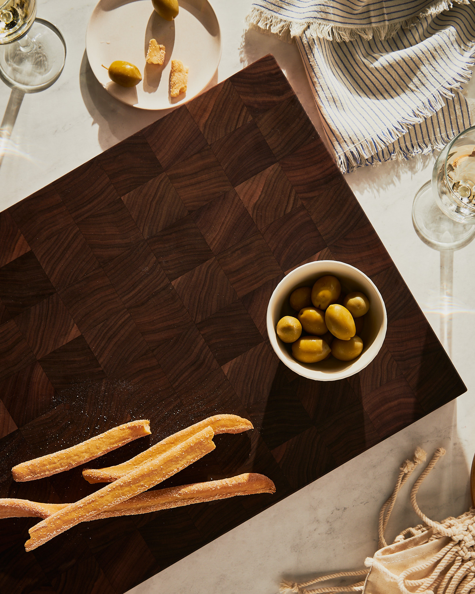 Vue en surplomb d'une planche à découper en bois de bout foncé garnie d'olives, de grissinis et de verres de vin au soleil, par Kata Endrodi, photographe culinaire à Montréal.