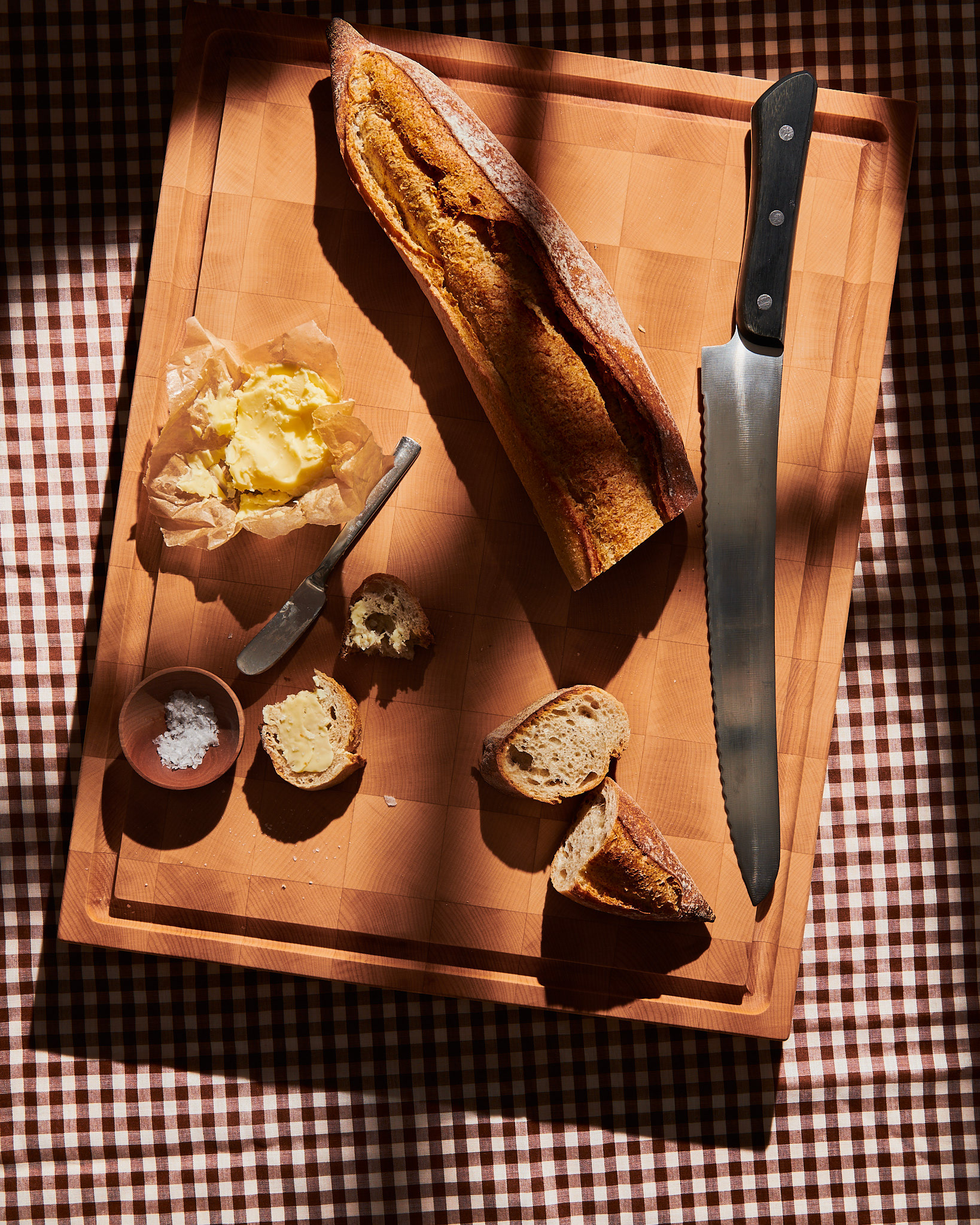 Vue en surplomb d'une baguette artisanale tranchée avec du beurre frais, de la fleur de sel et un couteau à pain sur une planche à découper au-dessus d'une nappe à carreaux, par Kata Endrodi, photographe culinaire à Montréal.