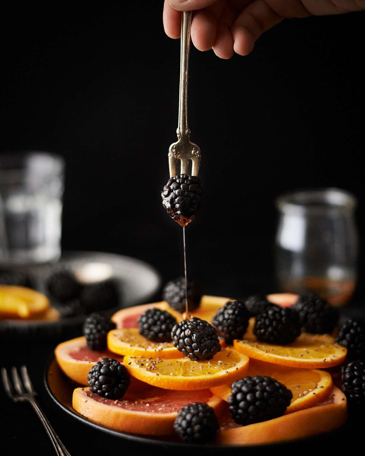 A food photography citrus plate image with blackberries, maple syrup and a hand holds a fork with a blackberry above plate, made by Kata Endrodi, a food photographer in Montreal