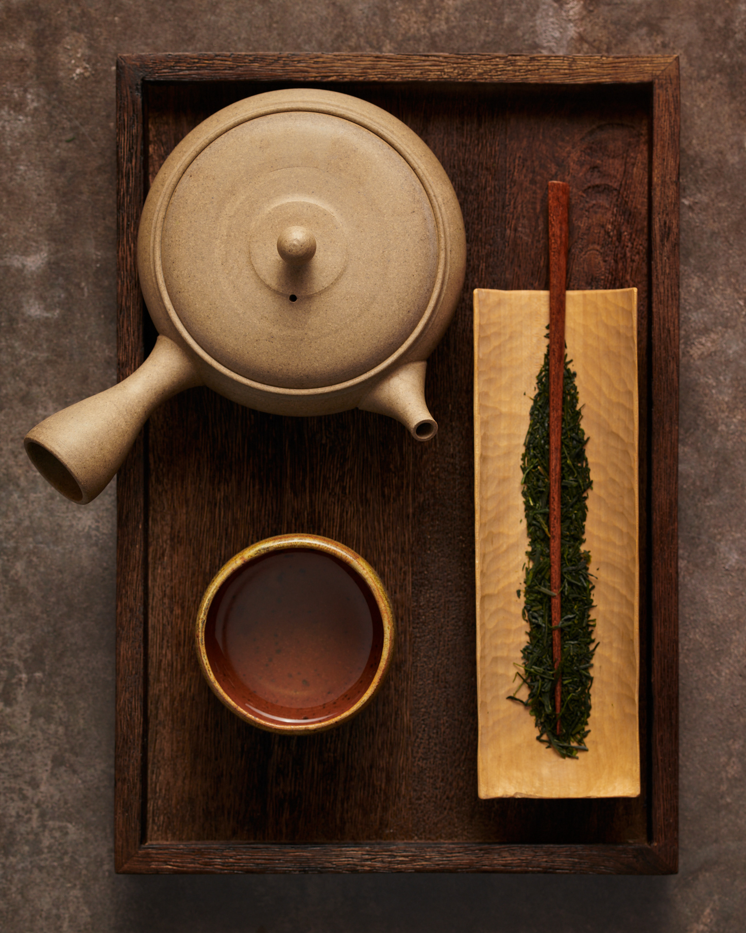 Kata Endrodi Montreal food and product photographer, top-down view of a minimalist tea ceremony setup featuring a clay kyusu teapot, a cup of steeped tea, and loose green tea leaves on a hand-carved wooden tray.