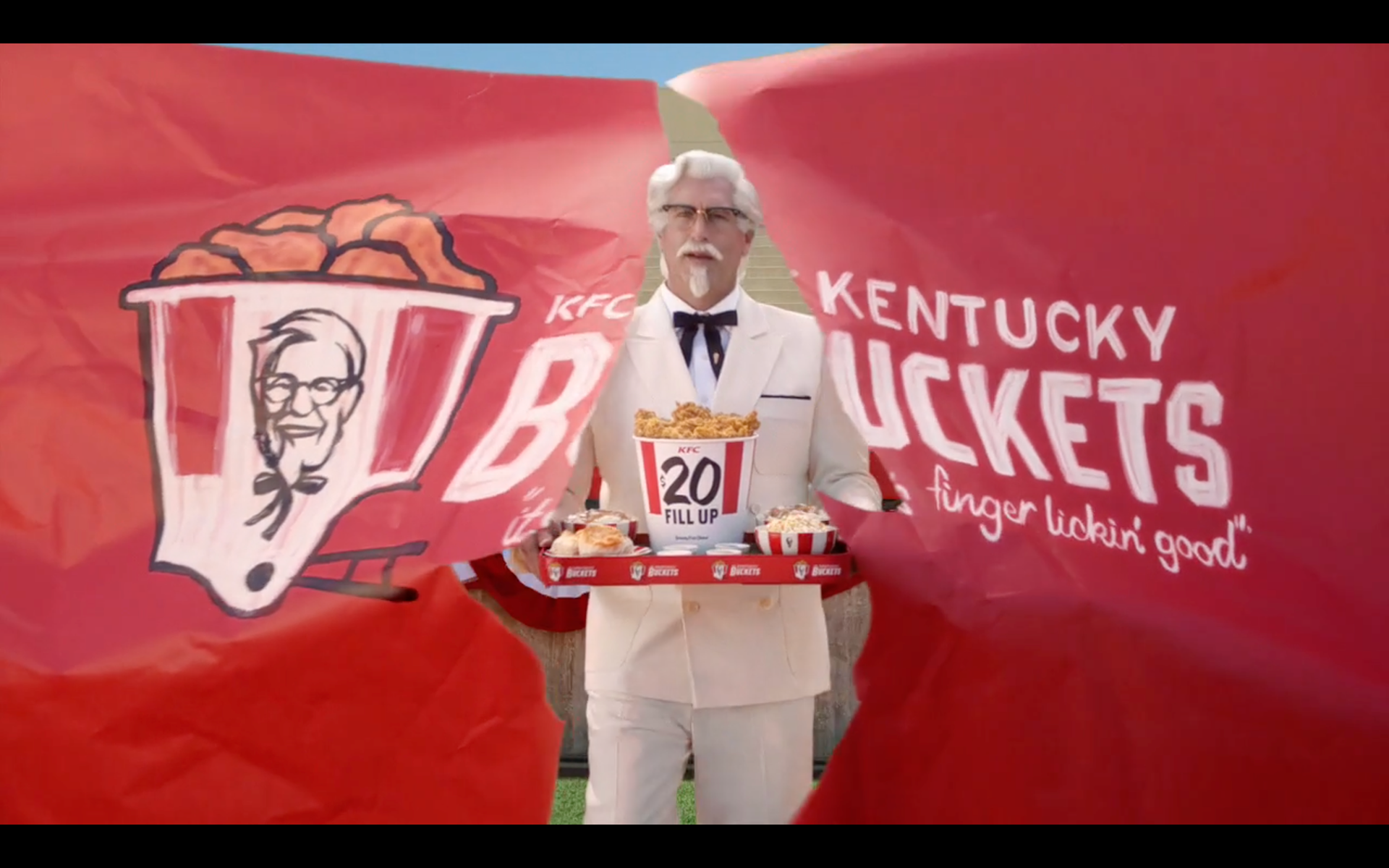 screenshot of KFC broadcast commercial introducing KFC Buckets starring Rob Riggle, who is bursting through a red kfc sign holding a platter of fried chicken.