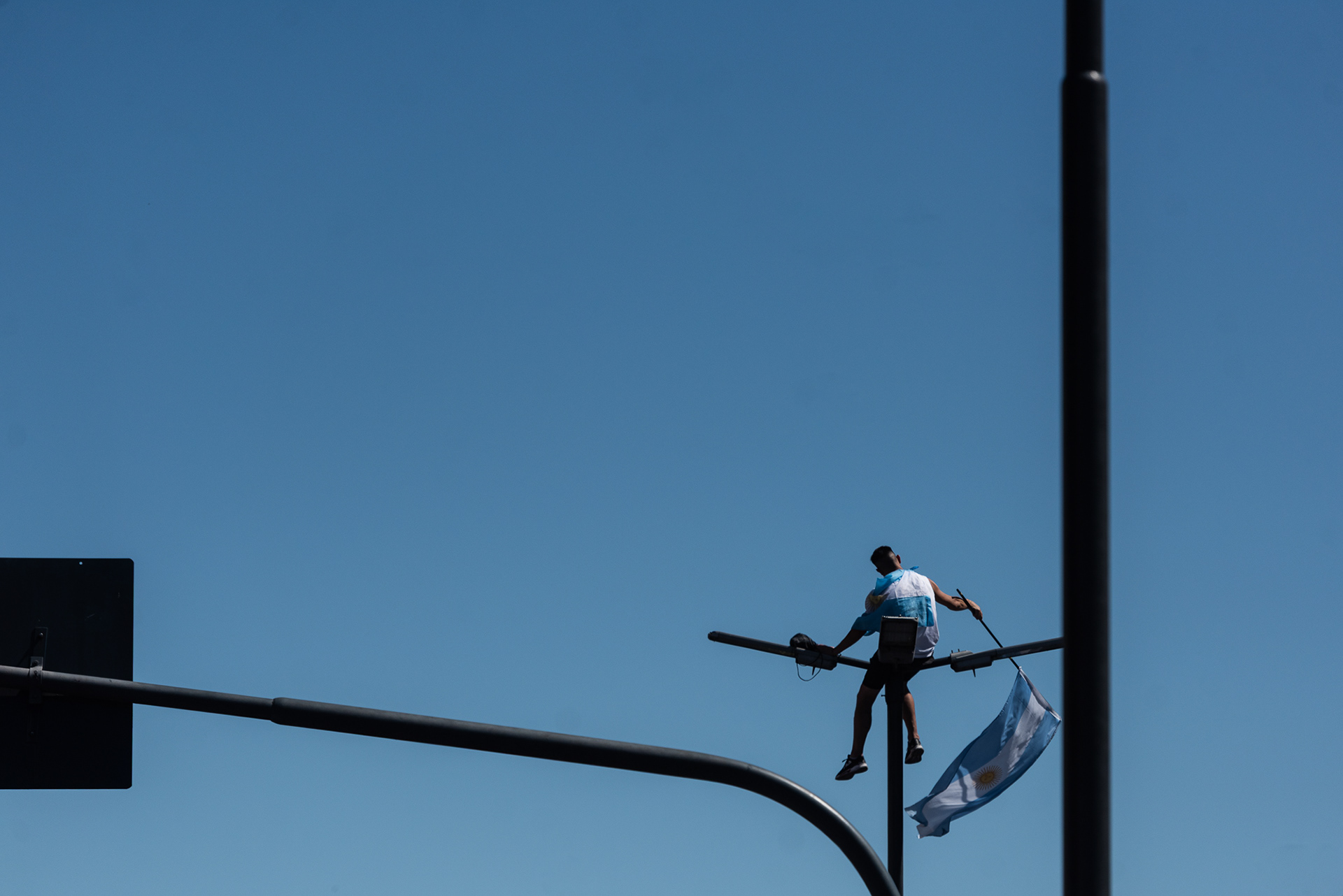 Elevado sobre el delirio, un hincha solitario conquista la altura, sentado sobre la luminaria pública, convierte el poste en un mástil improvisado para que el pabellón nacional se desgarre en el viento de la victoria.