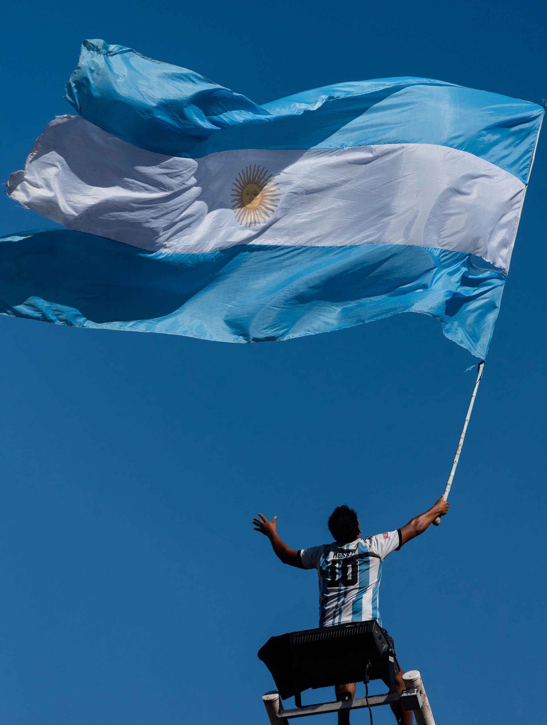 Con el rostro bañado en luz, un hincha alza la mirada al infinito, ofreciendo el furor de la bandera argentina al cielo como un voto de gratitud.