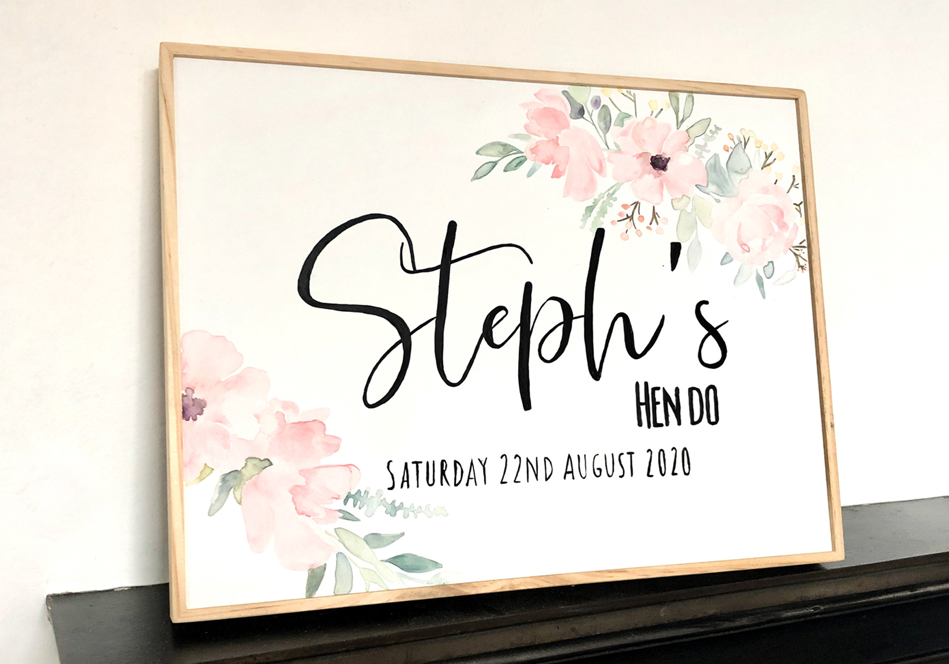 personalised "hen do board". a white canvas with a fine light wooden frame. on the canvas is the words "Steph'd HEN DO" and the date written in black & in the background is a water colour pink floral image in the bottom left and top right.