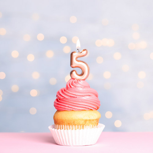 a cupcake with pink swirling frosting has a rose gold 5 candle lit resting on top. It is sitting on a pink surface and has a pale blue background with out of focus twinkling lights.