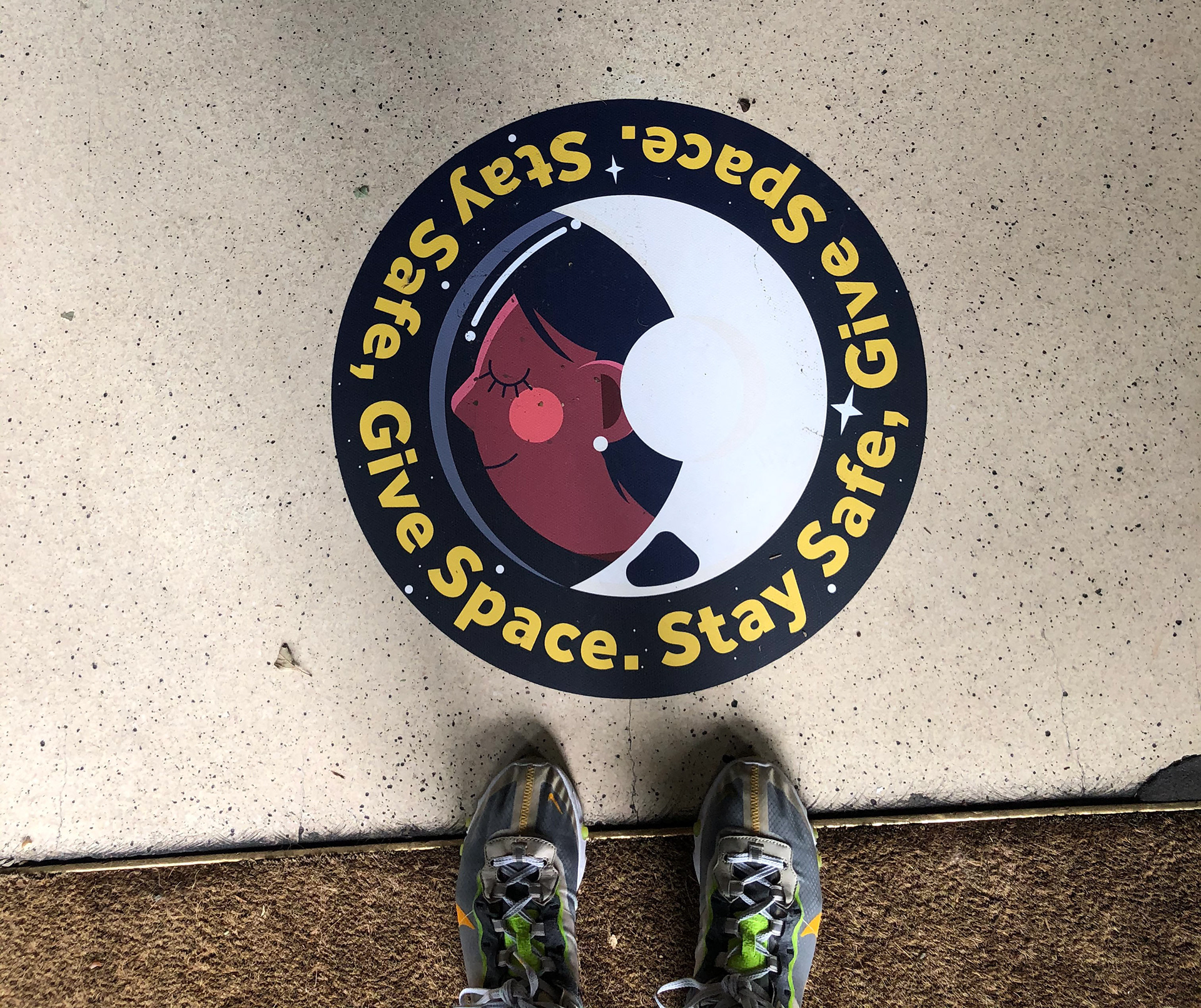 Circular floor decal showing a woman's profile in a crescent moon shape with text reading 'Space. Give Space. Stay Safe.' and a person's feet visible at the bottom, used for social distancing guidance.