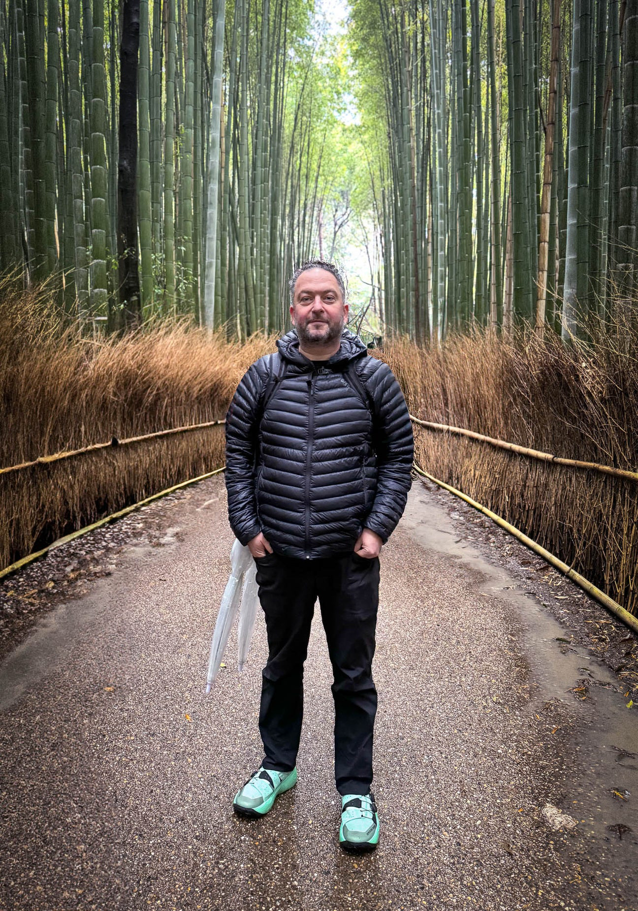 Person standing on a pathway in the Arashiyama Bamboo Grove in Kyoto, Japan. Tall green bamboo stalks tower on both sides, with bundled brush lining the path. The person is wearing a black puffer jacket, black pants, and mint green sneakers, holding a clear umbrella.