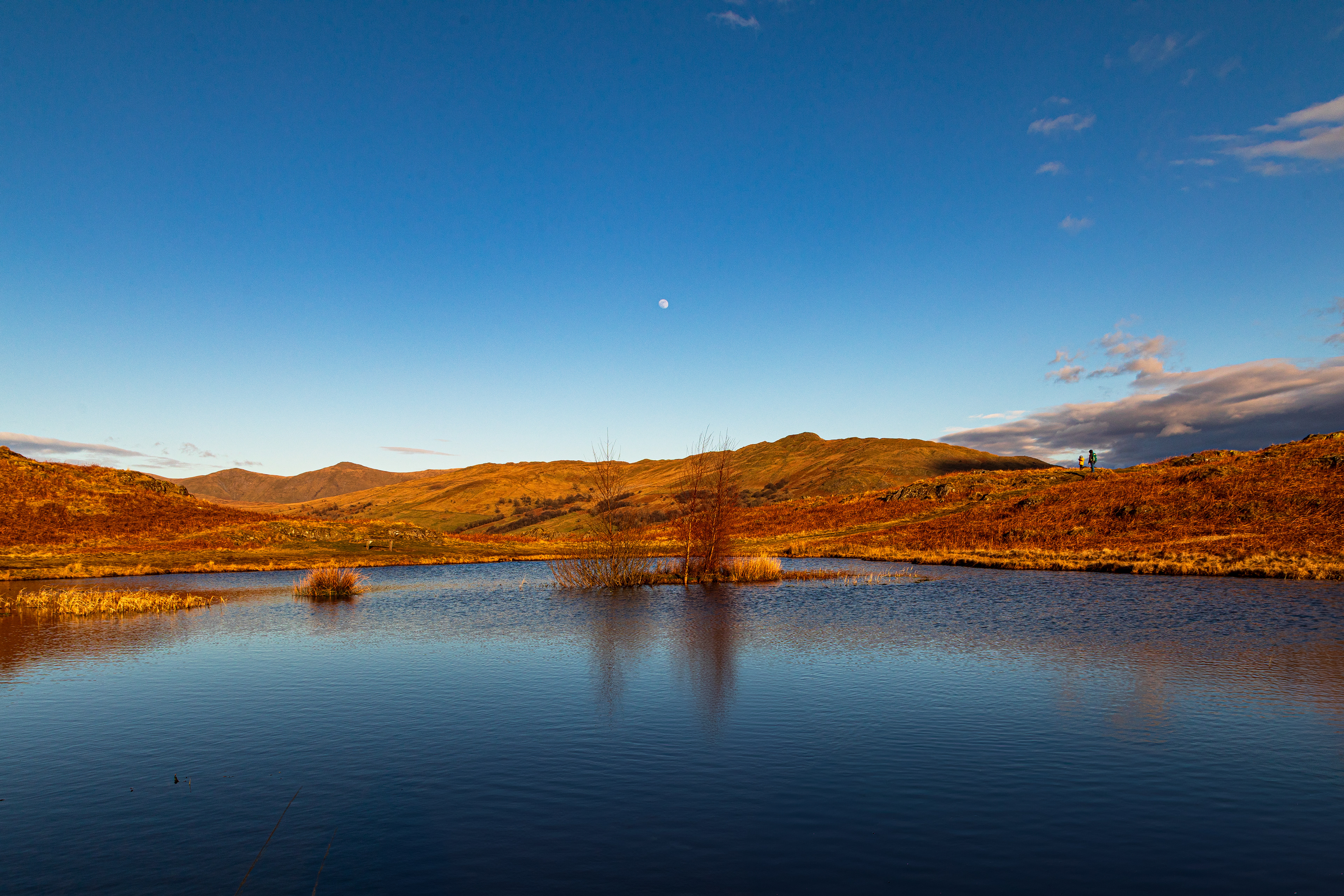 A small lake surrounded by hills with the moon above it