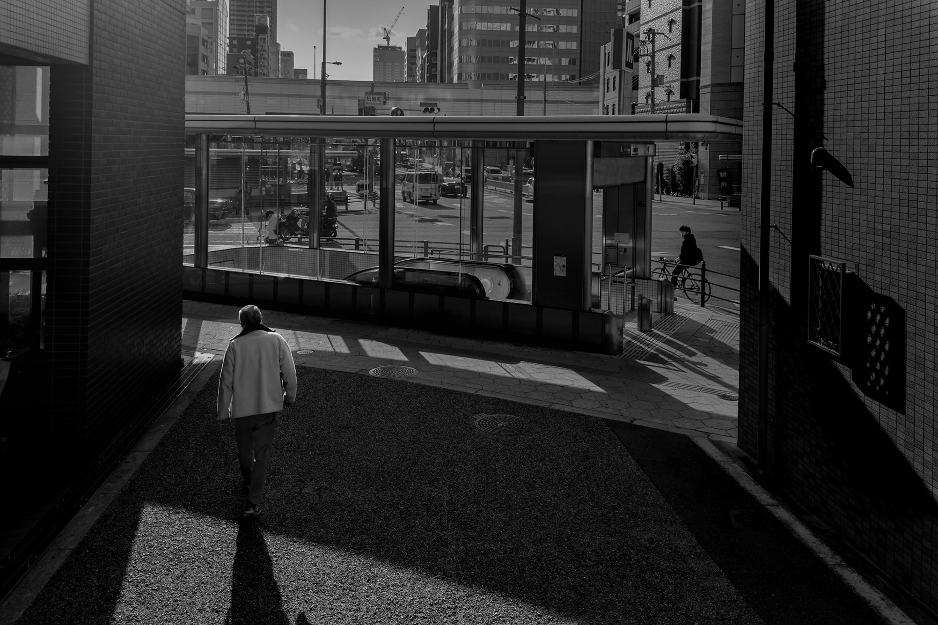 An elderly man walks through a sunlit passage in Osaka, his shadow the only thing keeping pace. 