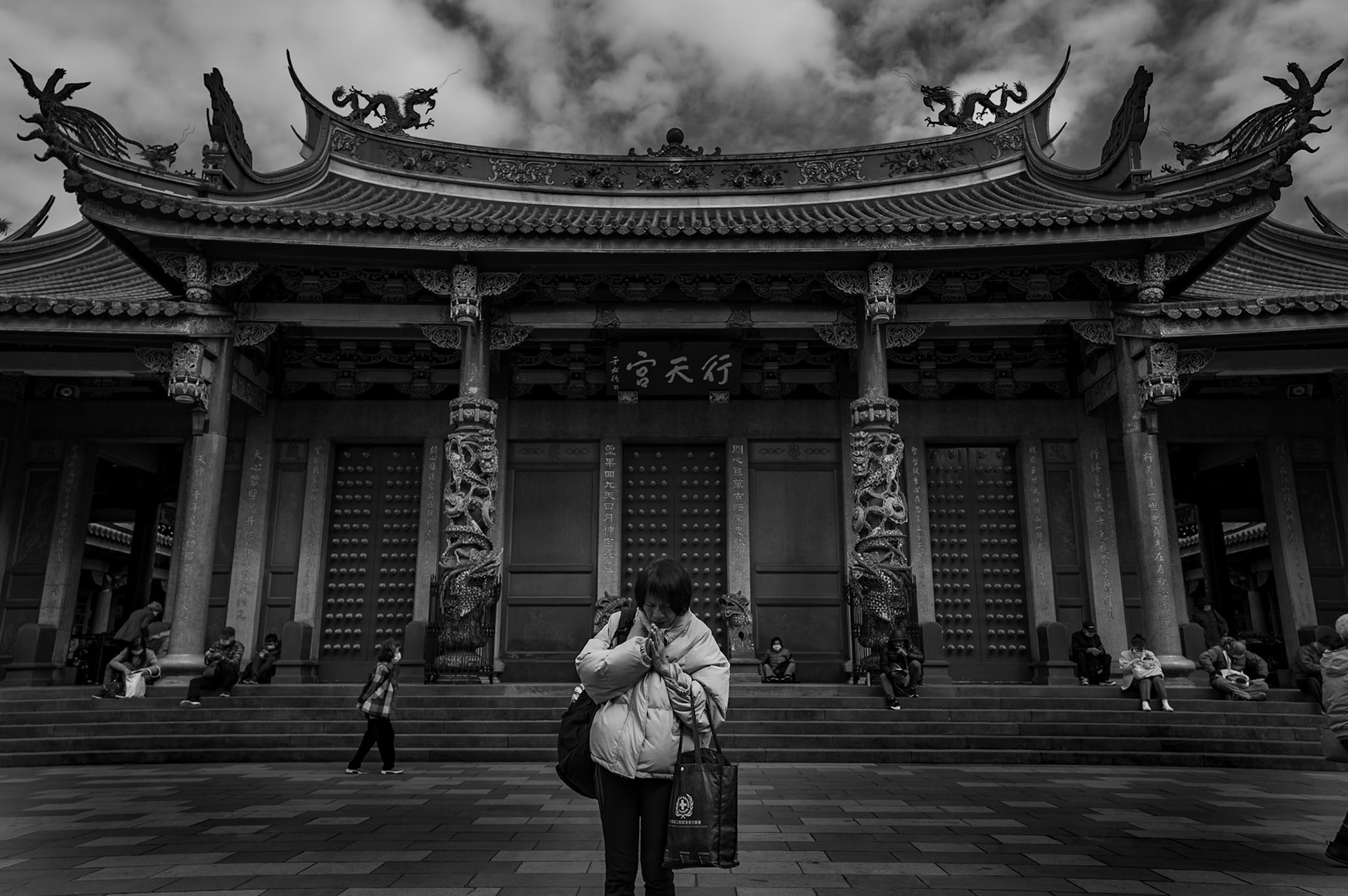 A woman prays alone in front of Xingtian Temple, turned inward, the crowd around her falling away.