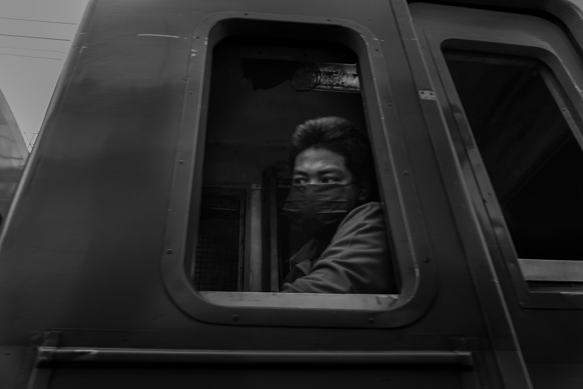 A man peers through the window of a train outside Bangkok, his face half-covered, eyes outward.