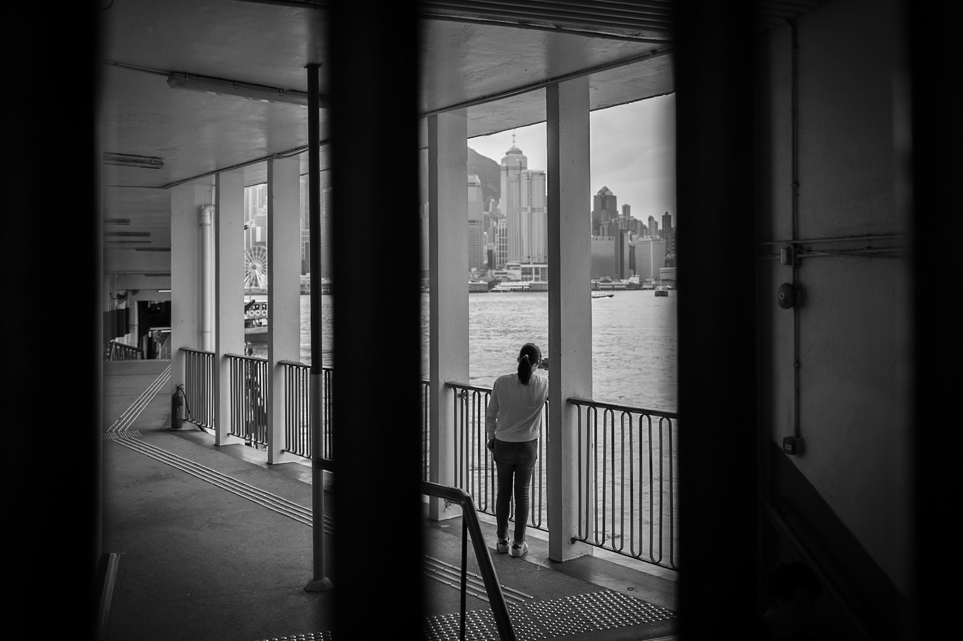 A woman stands alone at the railing of the ferry terminal, the Hong Kong skyline spread before her. 