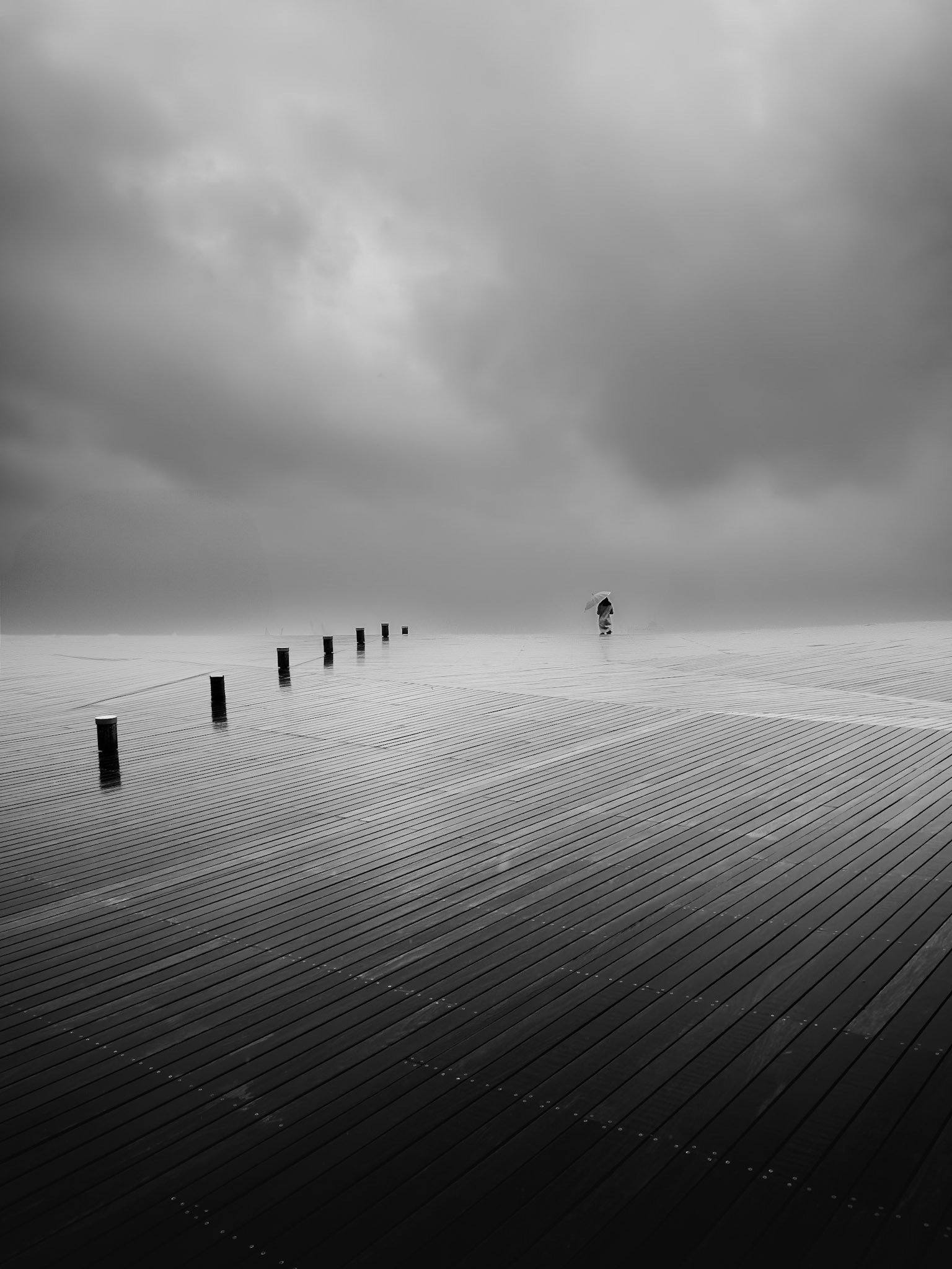 A lone figure stands on a rain soaked pier in Yokohama, dwarfed by open sky and water, holding still against the weight of both. 