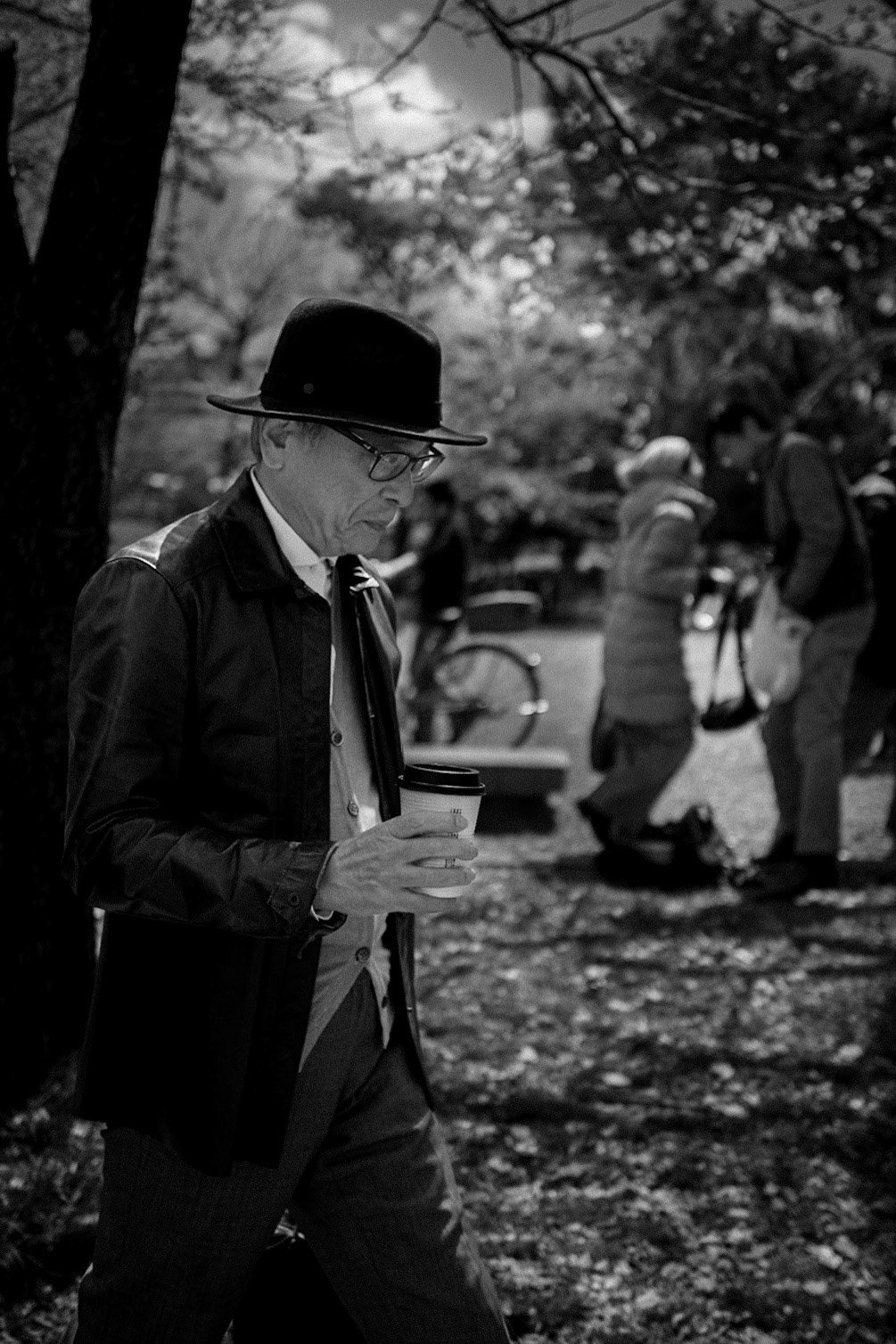 An elderly man pauses in Osaka Castle Park, coffee in hand, alone in thought while life continues around him.