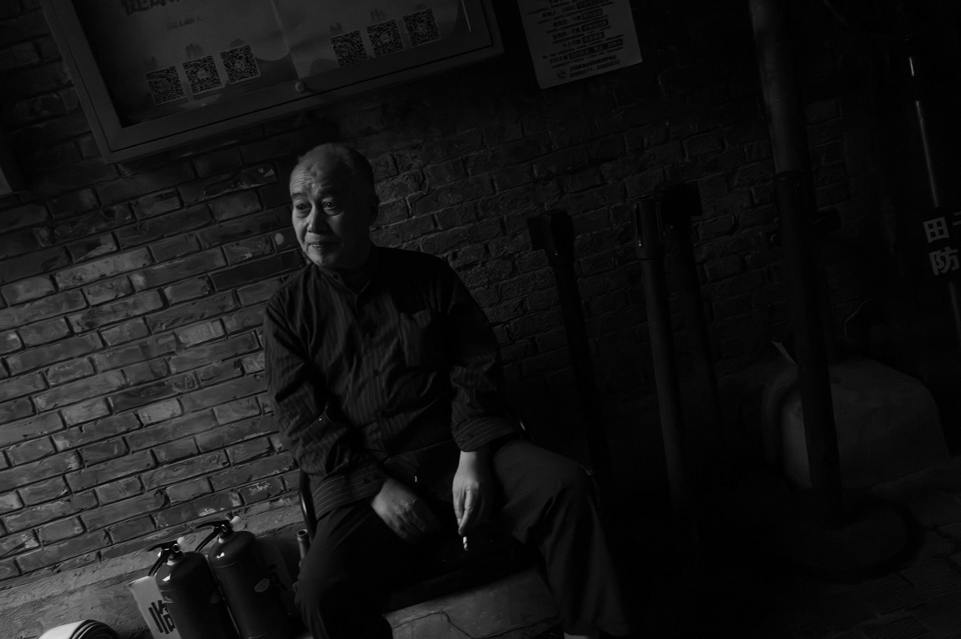 An elderly man sits alone against a brick wall, cigarette in hand, watching the world pass.